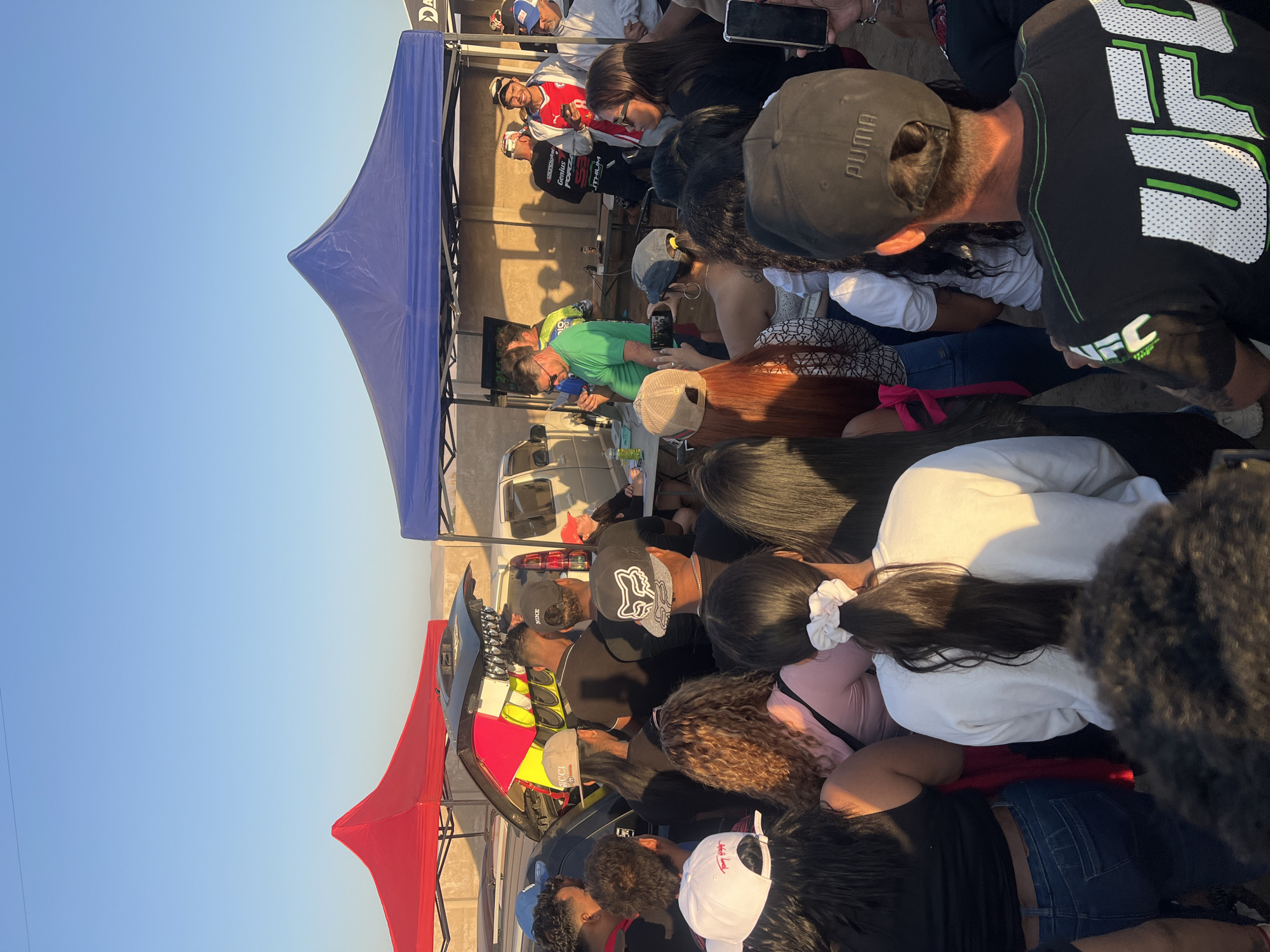A crowd of people surrounds a parked vehicle with an open trunk under a blue and red tent. People are looking at the contents of the trunk, where there is an object with multiple colors. Some are holding phones and recording. Behind the blue tent, there are people on a platform. The scene is outdoors in an open space with a clear blue sky. It could be in Arica, Chile, based on the lighting and the presence of a crowd gathered in a public area, but there is no explicit information in the image.