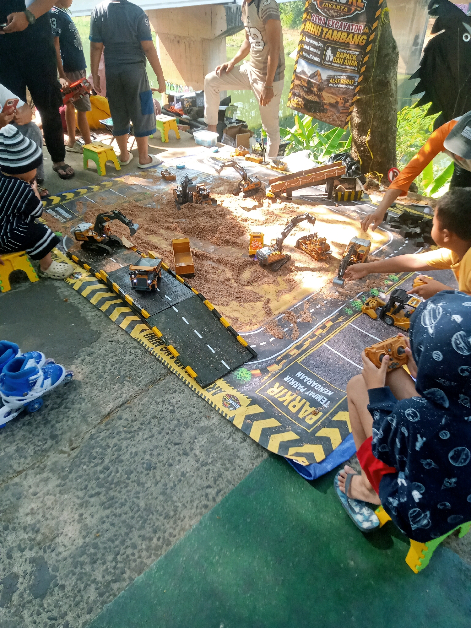 The image depicts an outdoor scene, likely in a park or public space, with several children engaged in playing with toy construction vehicles on a simulated construction site. The ground is a mix of concrete and a dark green mat. A large banner with Indonesian text, possibly advertising a toy excavation event, is visible in the background, attached to a tree. The children are interacting with the toy excavators, dump trucks, and other construction machinery spread across a mat depicting roads and a sandy area. One child is holding a remote control for a toy vehicle. The presence of a banner with "Jakarta" suggests the location is East Jakarta, Indonesia. The overall atmosphere appears to be recreational and lively.