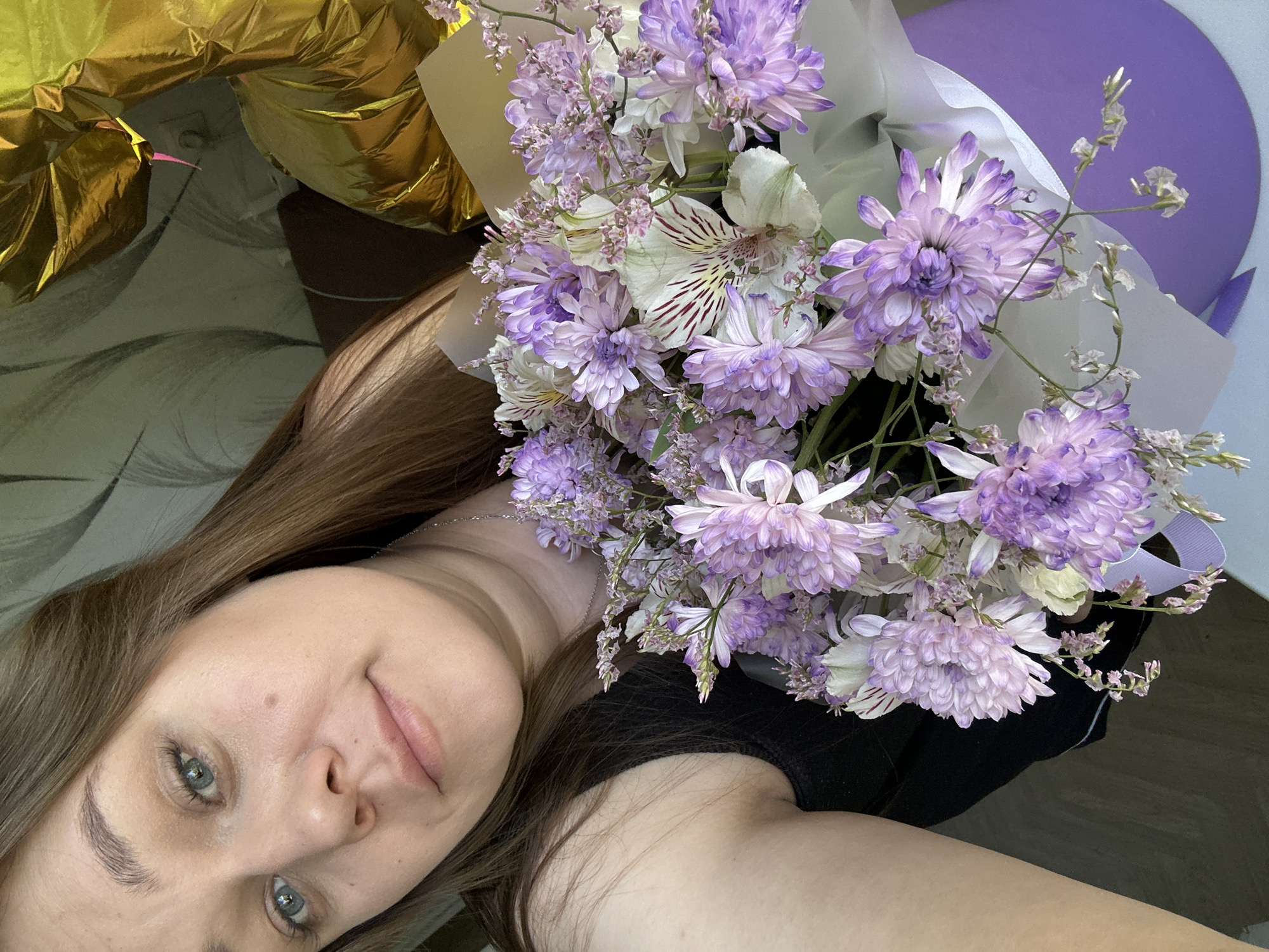 The image is a close-up selfie of a young woman lying down, taken from an overhead perspective. Her head is tilted to the left, showcasing her light brown hair, blue-green eyes, and a gentle, serene smile. She has clear skin and appears to be wearing light makeup. A subtle silver necklace is visible, and she wears a black top or strap.
Resting beside her is a large, vibrant bouquet of fresh flowers. The arrangement primarily features fluffy light purple chrysanthemums, white alstroemeria with distinctive reddish-brown striped centers, and delicate sprigs of light purple statice or similar filler flowers. The bouquet is elegantly wrapped in translucent white paper, backed by a solid lavender or light purple material.
In the upper left corner, partially visible and slightly out of focus, are several shiny golden metallic foil balloons, suggesting a celebratory occasion. The setting appears to be indoors, likely a casual home environment, with a patterned fabric visible behind her head. The lighting is soft and natural, indicating it is daytime. There is no visible text in the image. The scene conveys a relaxed and happy moment, possibly capturing the reception of a gift, in Chaikovsky, Russia.