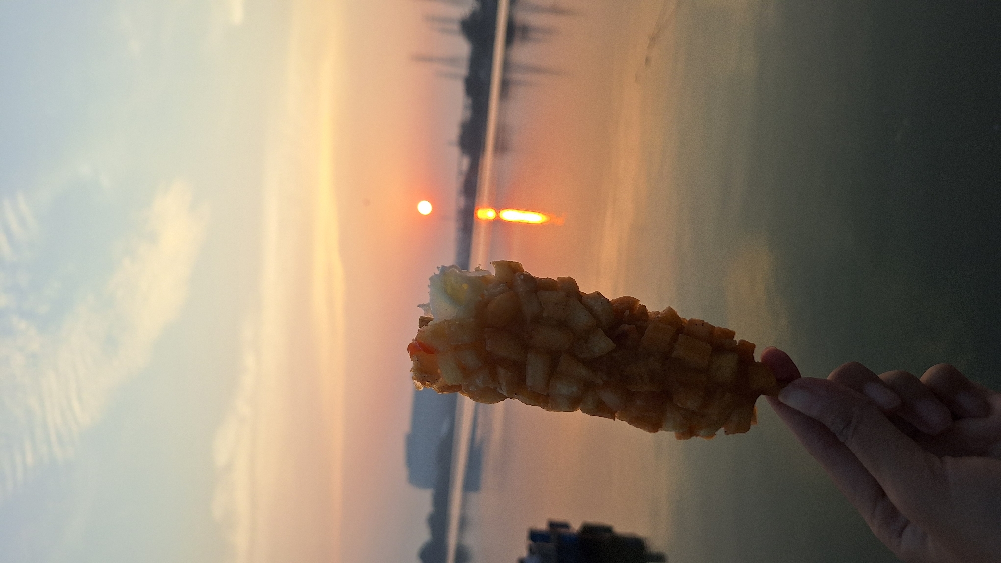 A hand holds a corn dog covered in cubed potatoes. The corn dog is in the foreground, slightly off-center to the left. Behind the corn dog, a body of water reflects the warm hues of a sunset. The sun itself is a small, bright orb positioned on the horizon. In the distance, silhouetted structures and cranes suggest an urban or industrial waterfront, possibly in North Jakarta, Indonesia, given the context. The sky is a soft blend of orange, pink, and blue, with wispy clouds.
