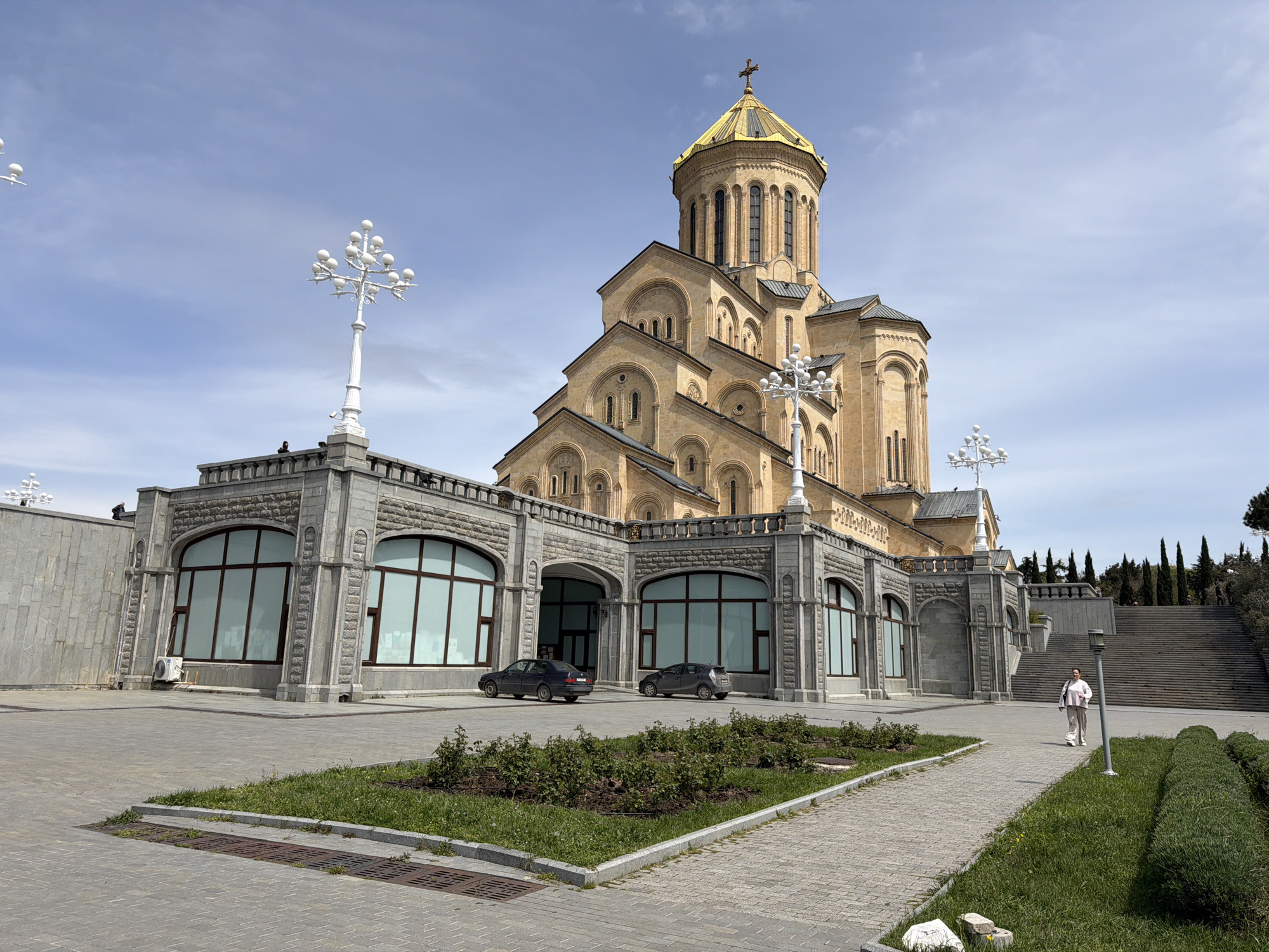 The image displays a wide view of a majestic Eastern Orthodox cathedral, likely the Holy Trinity Cathedral (Sameba) in Tbilisi, Georgia, under a bright, partly cloudy sky. The central church structure is built from light-colored stone, characterized by multiple tiers of arched windows and roofs, culminating in a striking golden dome topped with a cross.
In the foreground, a spacious, paved plaza extends, featuring landscaped sections with grass and small bushes. Flanking the main church are lower, multi-story buildings constructed from grey stone with large, arched windows and decorative stone carvings. Two dark-colored cars are parked in front of these lower structures. Several ornate, white lampposts with multiple globes illuminate the area, adding to the grandeur.
To the right of the frame, a woman in light-colored attire walks along a paved pathway, leading towards a wide stone staircase that ascends to an elevated area adorned with tall, slender evergreen trees. The scene is bathed in natural daylight, suggesting a clear, pleasant day. There is no visible text in the image.