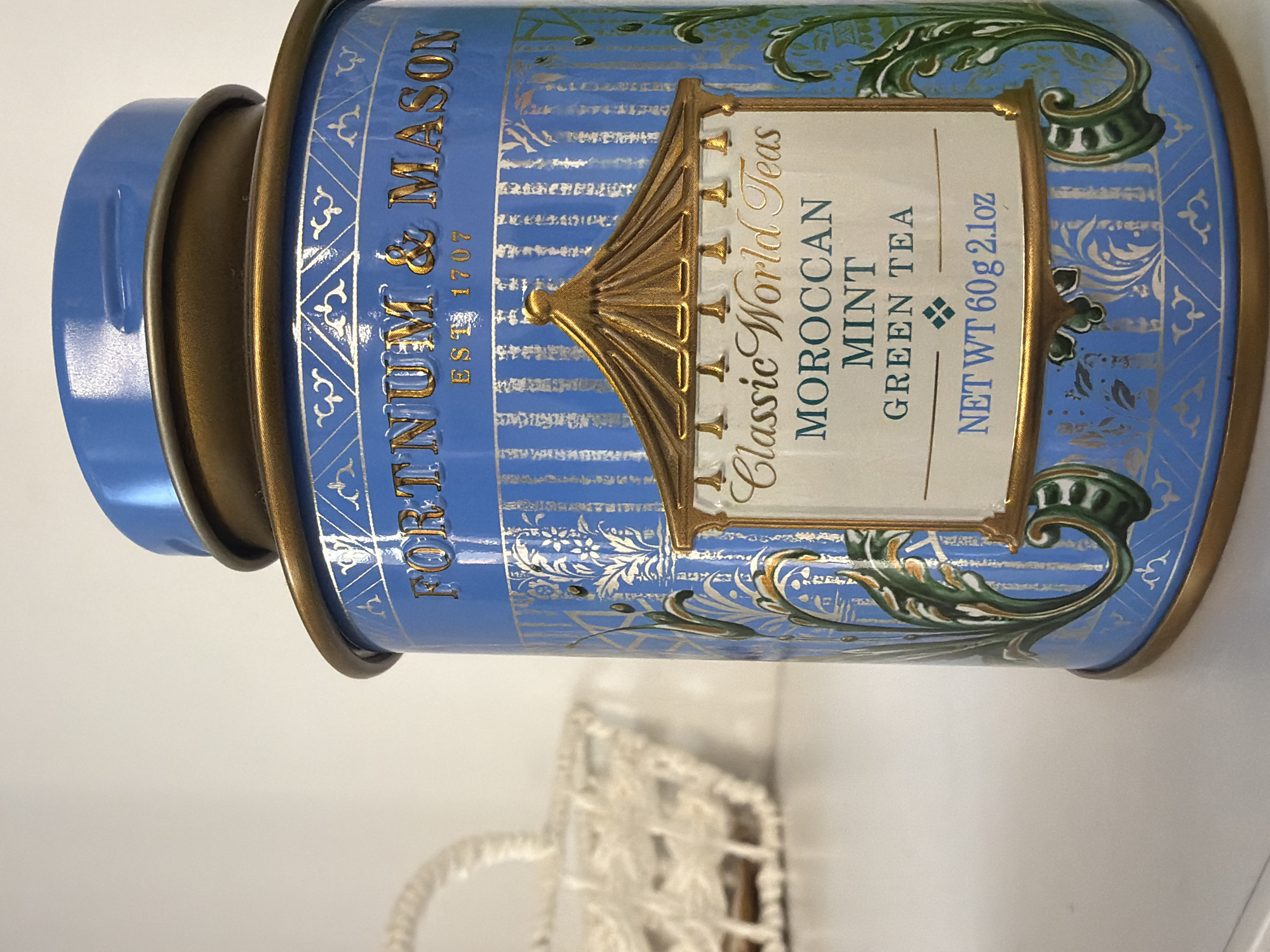 The image shows a blue tea canister with gold accents. It is labeled "FORTNUM & MASON EST 1707" and "Classic World Teas MOROCCAN MINT GREEN TEA NET WT 60g 2.1oz." The canister has a blue lid and gold bands. The image also includes part of a white woven object in the foreground. The location is not specified in the image.