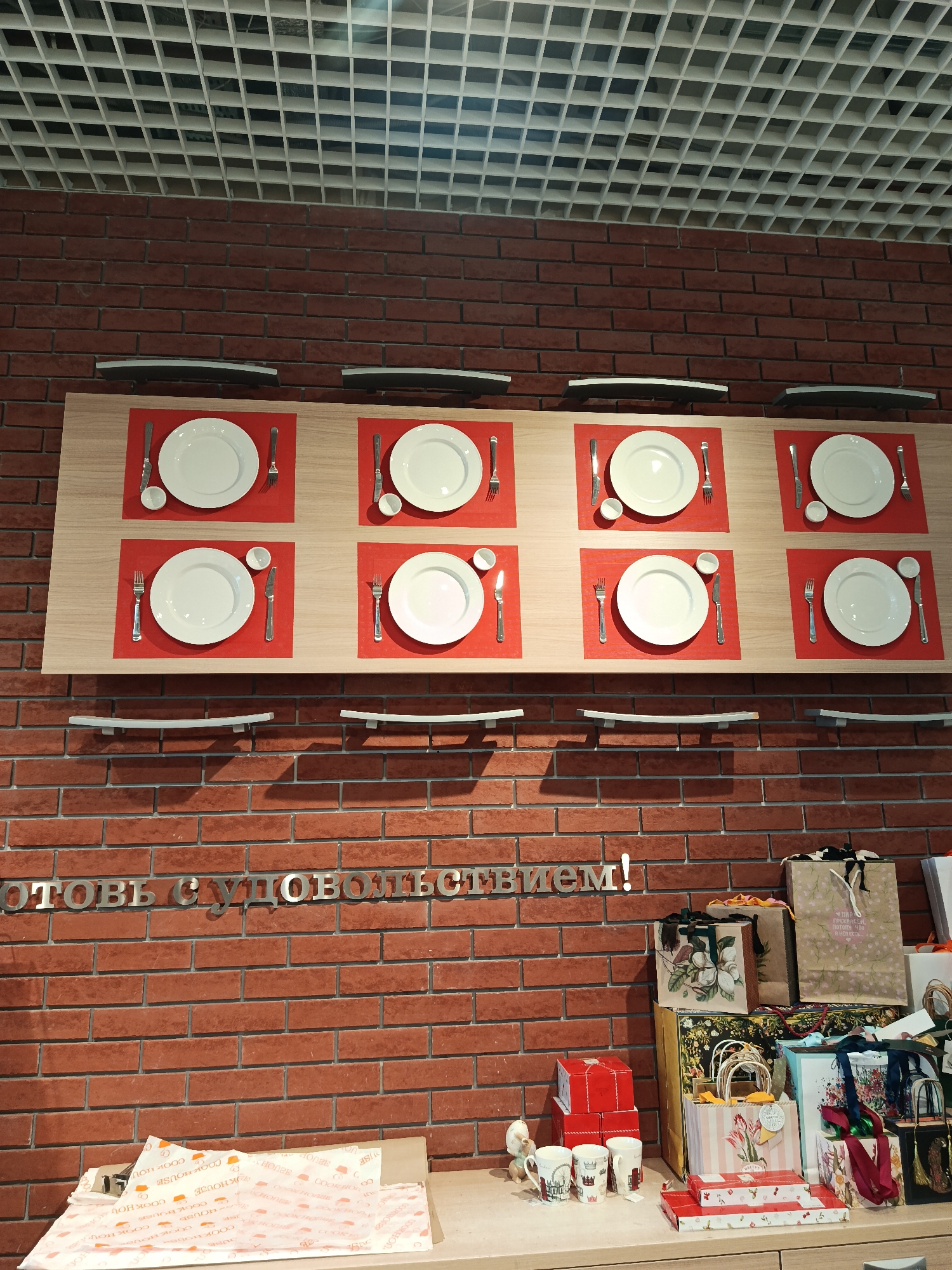 Here is a detailed description of the image:
**Scene:** The image appears to be taken inside a store or a restaurant, possibly in Moscow, Russia, as the Cyrillic text "Готовь с удовольствием!" translates to "Cook with pleasure!" in Russian. The scene is characterized by a combination of decorative elements and product displays.
**Environment:** The background is a red brick wall extending from the middle to the bottom of the shot, and the ceiling is covered in a white square grid.
**Objects:**
- At the center of the image, a set of six place settings (plates, silverware, and bowls) are displayed on a light wood panel attached to the brick wall. The place settings are arranged on red placemats.
- Below the light wood panel, there are two black shelves, seemingly decorative.
- The Cyrillic text "Готовь с удовольствием!" is attached to the brick wall and painted in silver.
- In the lower right corner, there is a display of merchandise, including shopping bags of various designs, red gift boxes stacked on top of one another, and mugs.
- A wooden countertop or table with a collection of items wrapped in paper with printed logos is visible in the bottom left corner.
**Subjects:**
- There are no people in the shot.
**Actions and Interactions:**
- The scene seems to be a curated display to showcase dining products or gift items.
