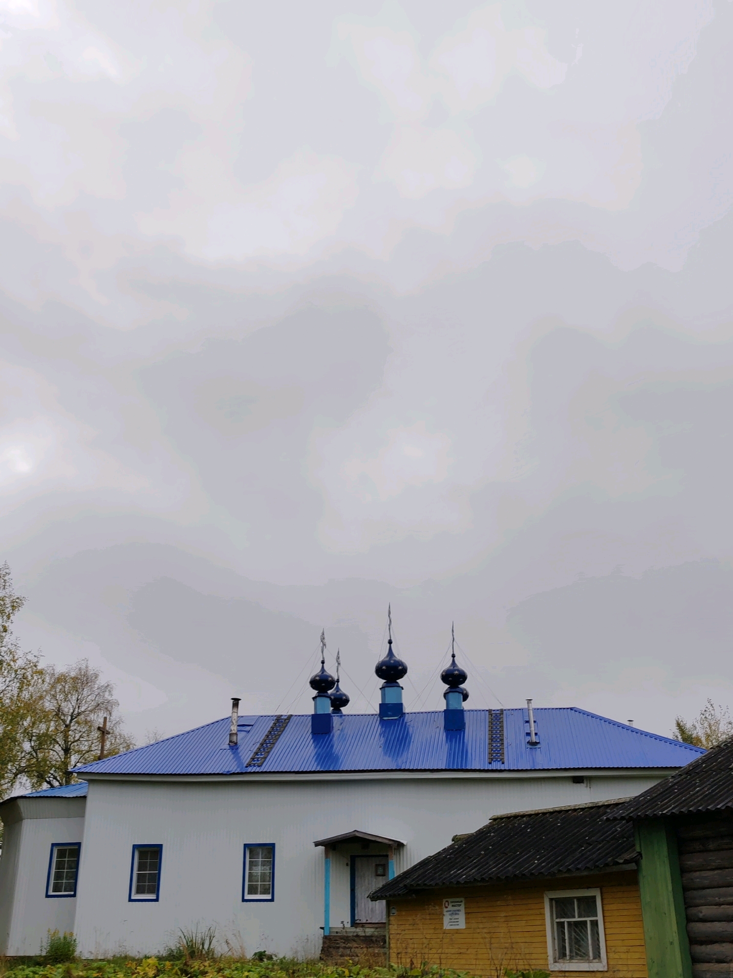 In the city of Chukhlem, Russia, a white building with a bright blue, ribbed metal roof stands under a cloudy sky. Five dark blue onion domes with spires and star-shaped finials are visible on the roof. The white building has several windows with blue trim. To the left of the main building, a smaller white structure is partially visible, also with a blue roof and a window. In front of the white building, a small, yellow, wooden outbuilding with a dark, corrugated roof is situated. A section of a dark-wood log building with a green trim is also present to the right. The ground in front of the buildings is covered with green foliage and some dry grass. Trees with yellowing leaves are visible to the far left. The overall scene depicts a rural or village setting, likely a church complex.