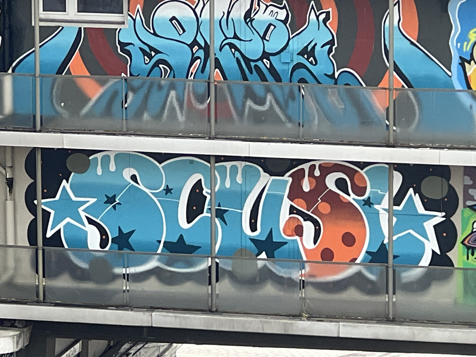The image shows a two-story building with graffiti art. The top level features blue and orange graffiti. The bottom level displays graffiti in blue and orange letters with star shapes. The building is located in Mönchengladbach, Germany.