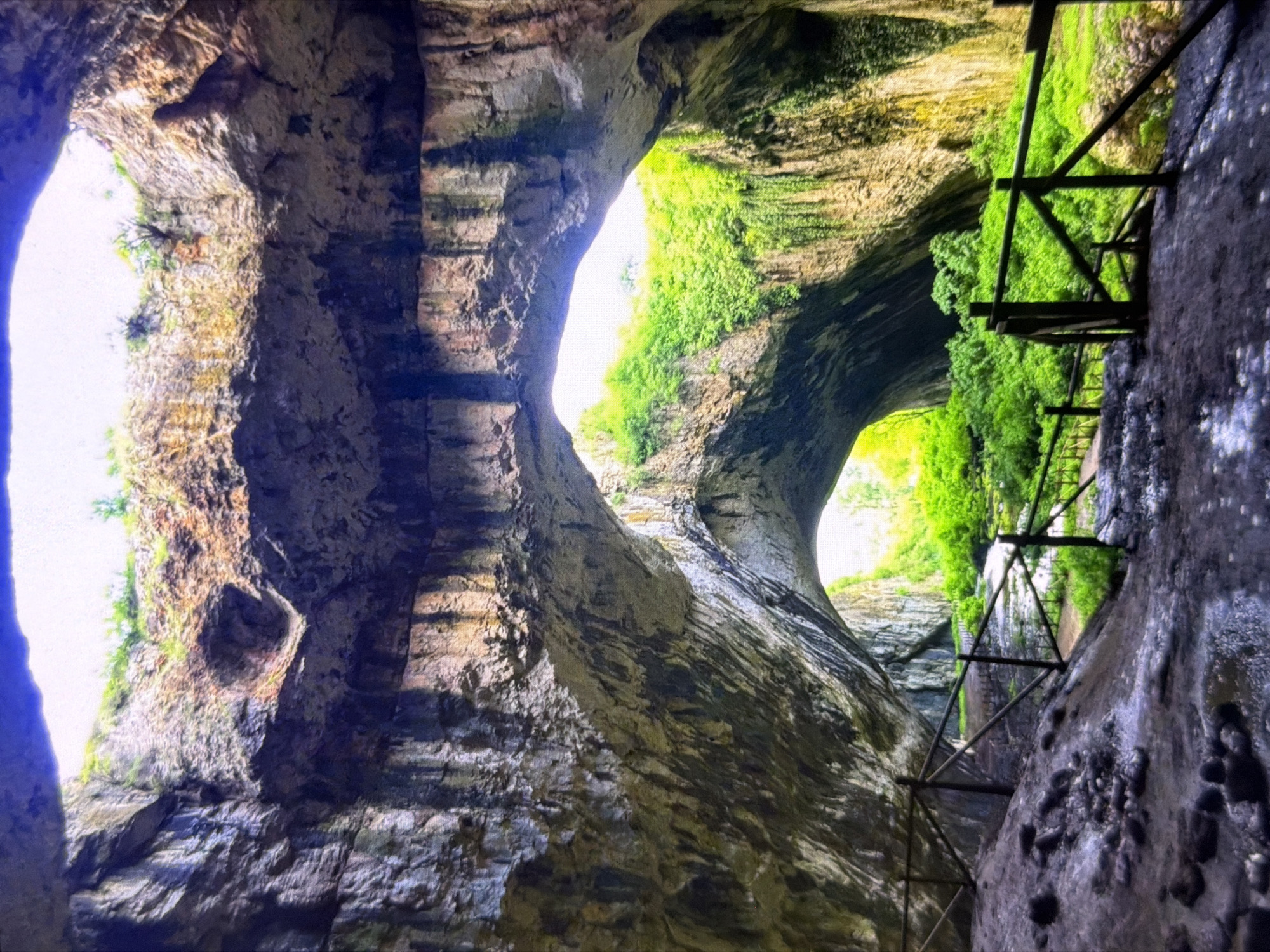 This image captures the majestic interior of a vast natural cave, likely the Devetashka Cave near Lovech, Bulgaria, viewed from within. The scene is dominated by immense rock formations, forming several massive arches and openings to the outside, allowing abundant natural daylight to flood into the cavern.
Bright green vegetation, including moss and small plants, thrives on the sun-kissed sections of the cave walls and hangs from the openings, contrasting sharply with the rugged, textured rock. The rock itself displays a range of colors from dark grays and browns in the shadowed areas to lighter tan and ochre where the light hits directly, revealing layers and striations.
On the right side of the frame, a man-made wooden structure, appearing to be a walkway, railing, or support, is visible, extending into the illuminated parts of the cave amidst the lush greenery. This structure suggests the cave is accessible to visitors. The strong light indicates it is daytime. No people or visible text are present in the image, emphasizing the natural grandeur and scale of the cave.