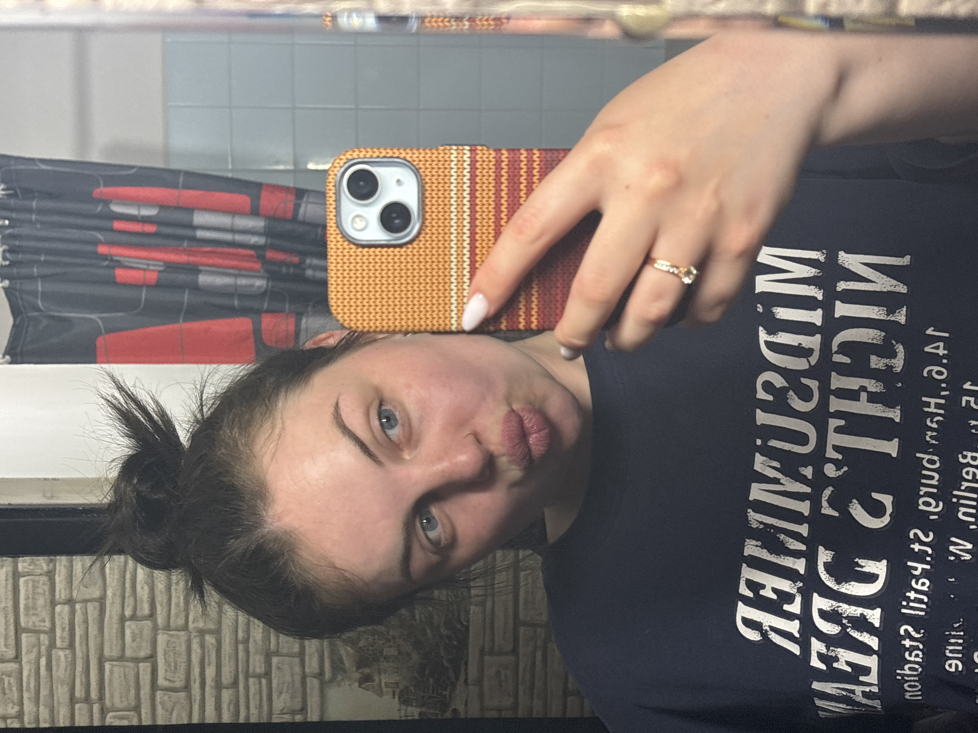 A woman is taking a selfie in a bathroom mirror, her hair in a messy bun. She is wearing a dark t-shirt with silver lettering. Her phone has a colorful knitted-style case. A ring is visible on her finger. The bathroom has a shower curtain with a graphic design and a tile wall. The image does not reveal any information to determine if the location is in Chornomorskoe, Ukraine.