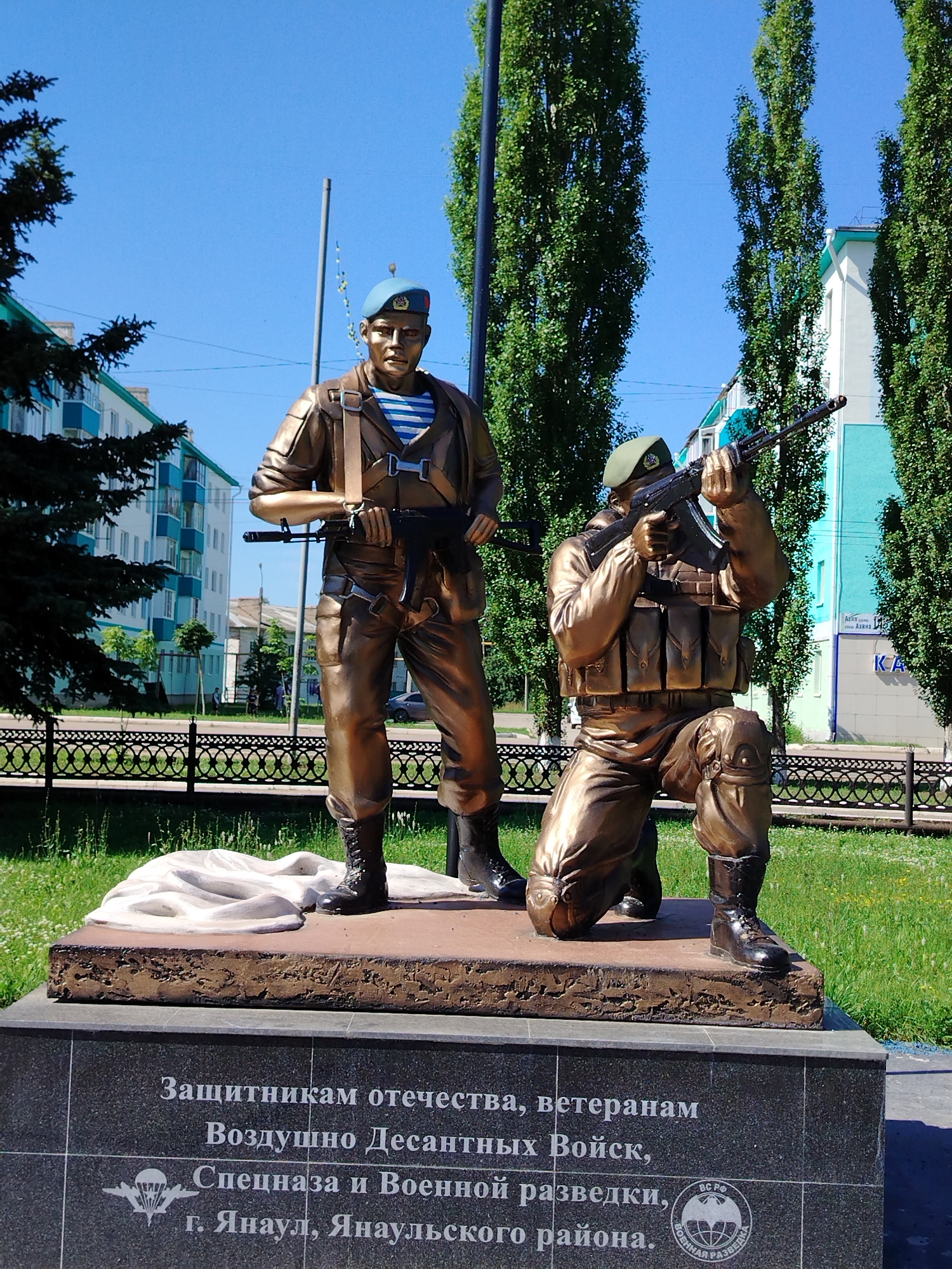 This is a photograph of a bronze monument depicting two paratroopers. The paratrooper on the left stands upright, wearing a blue beret, a striped undershirt, a military jacket, and trousers. He holds a rifle in his hands. The paratrooper on the right kneels, wearing a green beret, a tactical vest, and trousers, aiming a rifle. Both figures are detailed and appear to be in action poses.
The monument is situated on a rectangular stone base, which sits atop a larger granite pedestal. The granite pedestal bears an inscription in Russian, which translates to "To the defenders of the fatherland, to the veterans of Airborne Troops, Special Forces and Military Intelligence. Yanaul, Yanaulsky District." The inscription is accompanied by a circular emblem depicting wings and a globe, along with the words "ВОЕННАЯ РАЗВЕДКА" (Military Intelligence) and "ВС РФ" (Armed Forces of the Russian Federation).
The monument is located outdoors, in what appears to be a park or public square. The background includes tall, green trees, a light blue sky, and buildings with light-colored facades and turquoise accents. A black wrought-iron fence runs along the perimeter of the monument's area. Green grass covers the ground around the monument. The overall setting suggests a solemn and commemorative public space in the city of Yanaul, Russia.