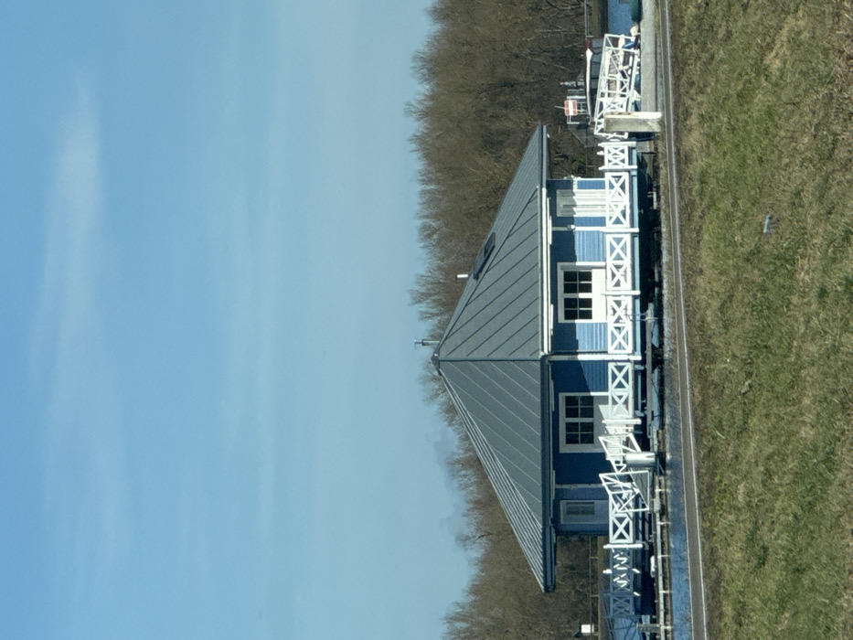 The image displays a bright blue, single-story house with a gray pyramidal metal-seamed roof situated on a body of water in Altenheim, Germany. The house features white exterior trim, multiple white-framed windows, and white doors, some appearing as double French doors, all surrounded by a white railing with a repeating 'X' pattern, forming an elevated deck or porch. A small vent/chimney and a skylight are visible on the roof. The structure appears to be supported by a submerged or floating platform.
To the left of the house, additional white railings extend over the water. To the right, a white sloped gangway or ramp descends into the water, partially obscuring a white pontoon-style boat. A red-and-white striped pole stands vertically near the boat. The immediate foreground consists of a strip of green grass with a horizontal dirt path or road. The background is dominated by a dense line of bare deciduous trees. The sky above is clear and light blue with minimal cloud cover. No individuals are present in the scene, and no overt actions or interactions are occurring.