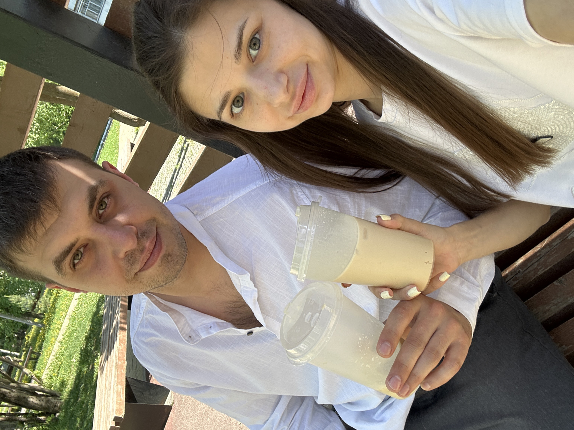 A man and a woman are posing for a selfie outdoors. The man is on the left, wearing a partially unbuttoned white collared shirt, while the woman is on the right, wearing a white t-shirt. They are holding two plastic cups with lids; one appears to contain a tan-colored liquid, and the other a white liquid. The background shows a wooden structure and a grassy area with trees, suggesting they are in a park or garden setting. The angle of the photo is slightly tilted. Based on the camera perspective and the presence of the wooden structure, it is likely that the photo was taken at the Izhevsk Zoo in Izhevsk, Russia, where the terrain could be elevated to a higher perspective.