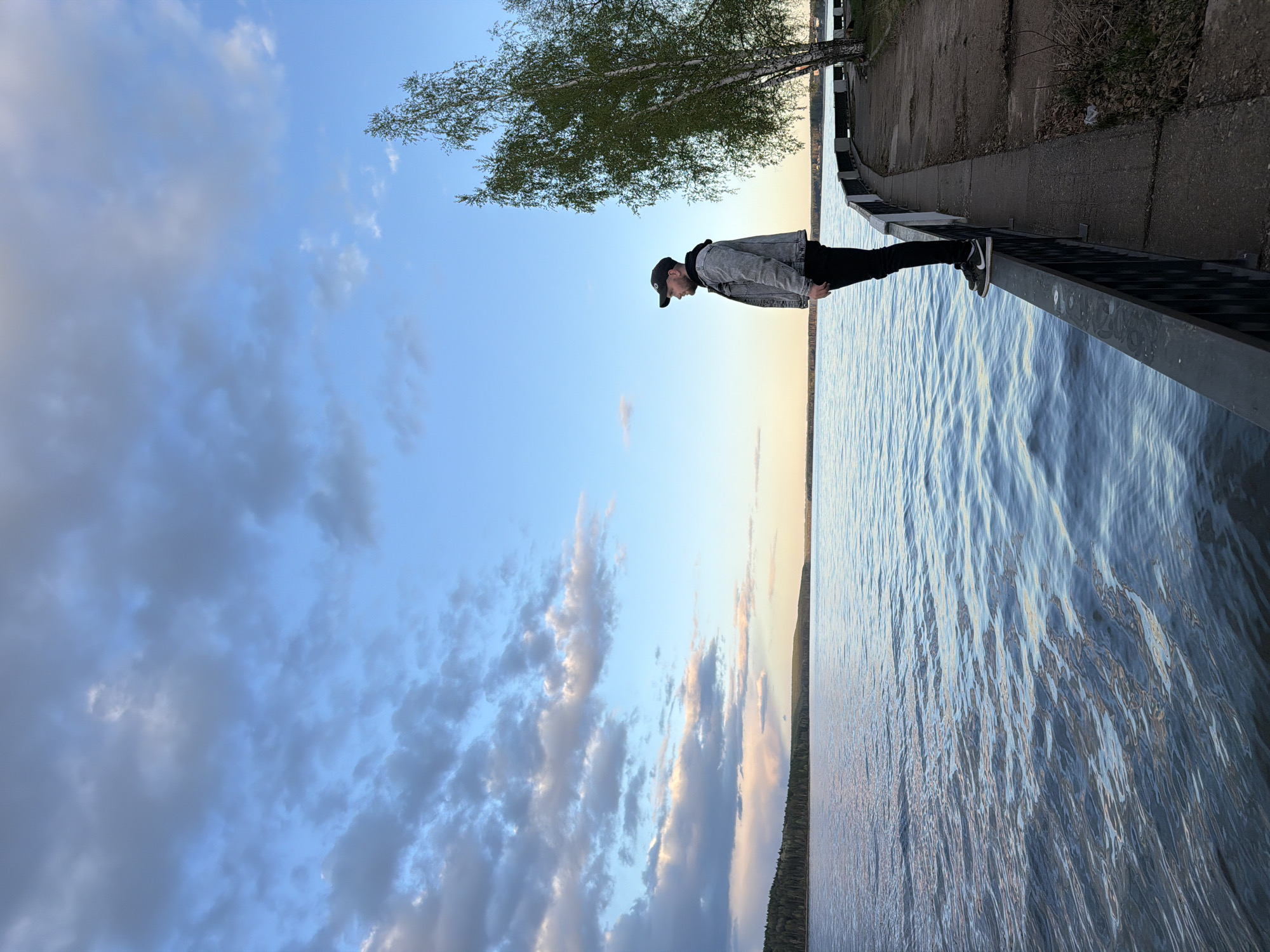 A person is standing on a bridge railing in Chaikovsky, Russia, with a body of water to their right and a sky with clouds to their left. The person is wearing a jacket, dark pants, and dark shoes, with a baseball cap on their head. The bridge has a concrete walkway and a metal railing. A tree is visible behind the person. The sky contains a mix of blue sky and greyish clouds, possibly reflecting the light of the setting sun. The water has ripples.