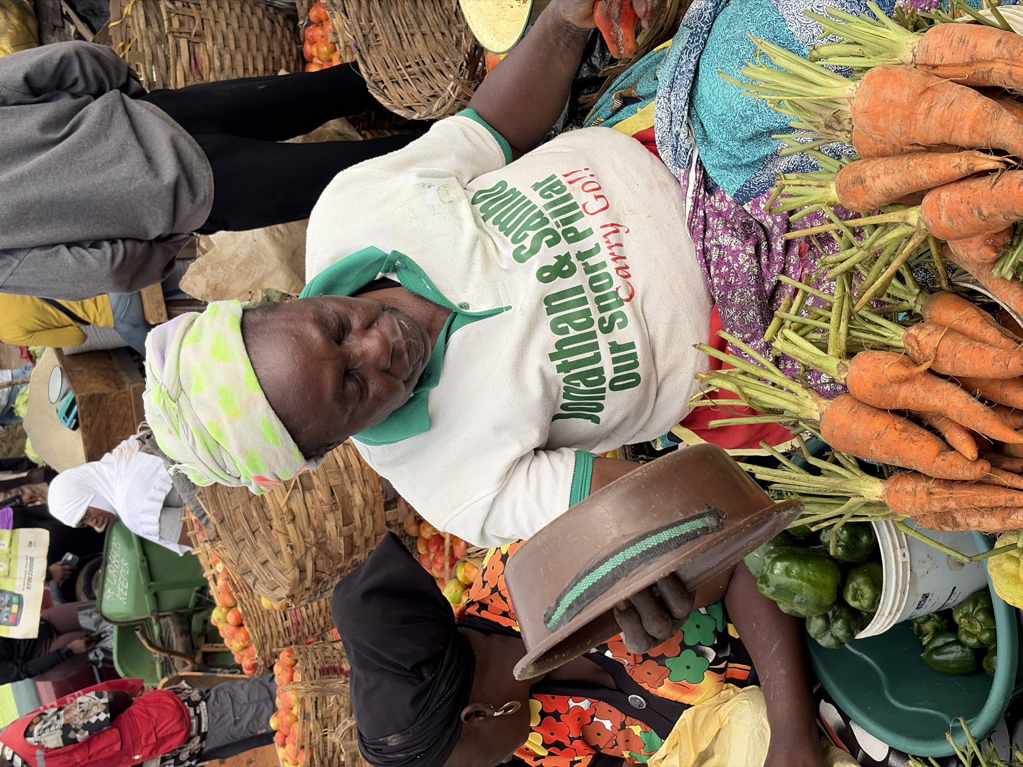 At a market in Gura Top, Nigeria, a woman wearing a white t-shirt with green trim and a green and white patterned headscarf is seated. Her t-shirt has text printed on it in green and red, which reads "Jonathan & Sambo Our Sport Pillar Carry Go!!". She holds a brown scoop in her left hand. In front of her, on the ground, are several green bell peppers in a turquoise bucket and a bunch of carrots with green tops. To her left, another woman in a black headscarf and a brightly colored floral shirt is partially visible. In the background, there are large woven baskets filled with red tomatoes, and other people are visible further in the background, including a person in a red vest and another in a yellow shirt. The setting appears to be an outdoor market stall with various produce and goods for sale.