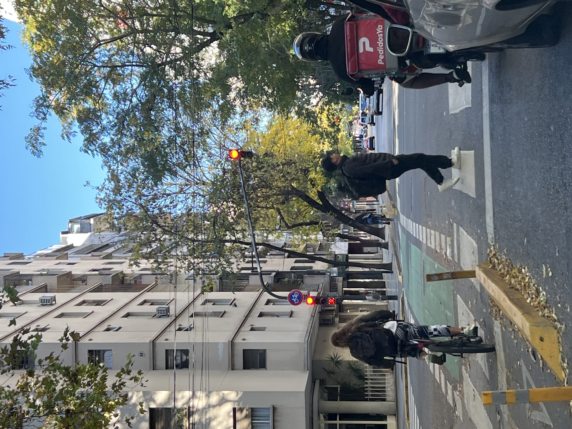 The scene is a street in Buenos Aires, Argentina. A person is crossing the street at a pedestrian crossing while the traffic light is red. A motorcycle with a delivery box labeled "PedidosYa" is stopped, presumably waiting for the light to change. Another person on a bicycle is stopped at a bike lane traffic light which is also red. A tall apartment building is on the left. Trees line the street, and the sky is visible in the upper left corner. The photo is oriented on its side.
