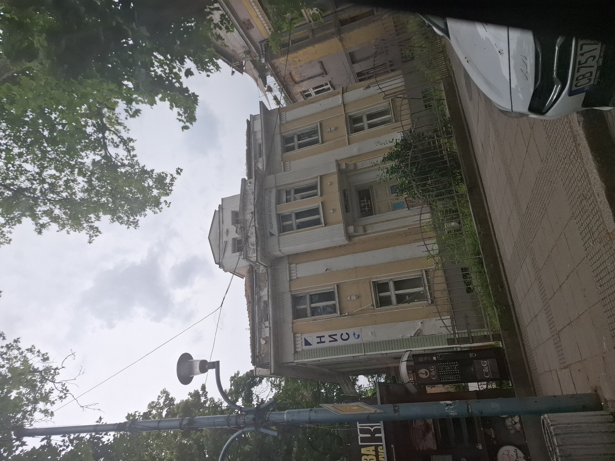 An eye-level, medium outdoor shot captures an urban scene under an overcast sky, focusing on an old, pale yellow and white building with peeling paint. The building features multiple windows, some with white shutters, and decorative architectural elements. To the left of the building, a lamppost stands tall, with a sign displaying Cyrillic text vertically on the building's facade. Adjacent to the building, another older, partially damaged building with yellow and grey tones is visible. A metal fence runs along the front of the buildings, separating them from a sidewalk paved with light brown square tiles. A white car with a license plate reading "CB 7517" is parked on the right side of the frame, partially obscuring the view. Trees with green foliage frame the top of the image.