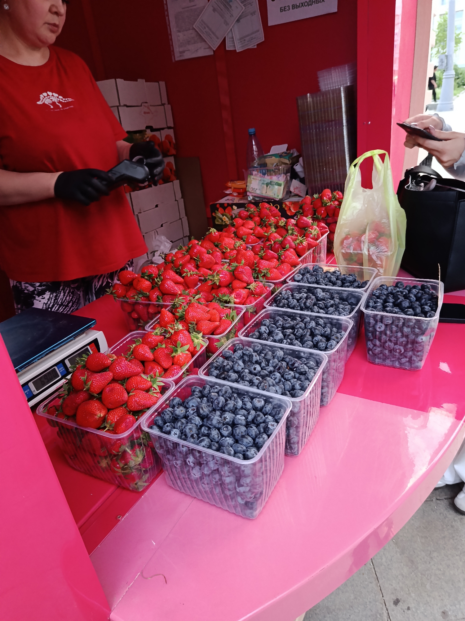 At an outdoor market in Moscow, Russia, a vendor wearing a red t-shirt and black gloves uses a card reader to process a transaction with a customer. The vendor is standing behind a pink table displaying containers of fresh strawberries and blueberries. Behind the vendor are stacked boxes of strawberries. A plastic bag filled with strawberries sits near the customer's black handbag. The market stall is predominantly red.