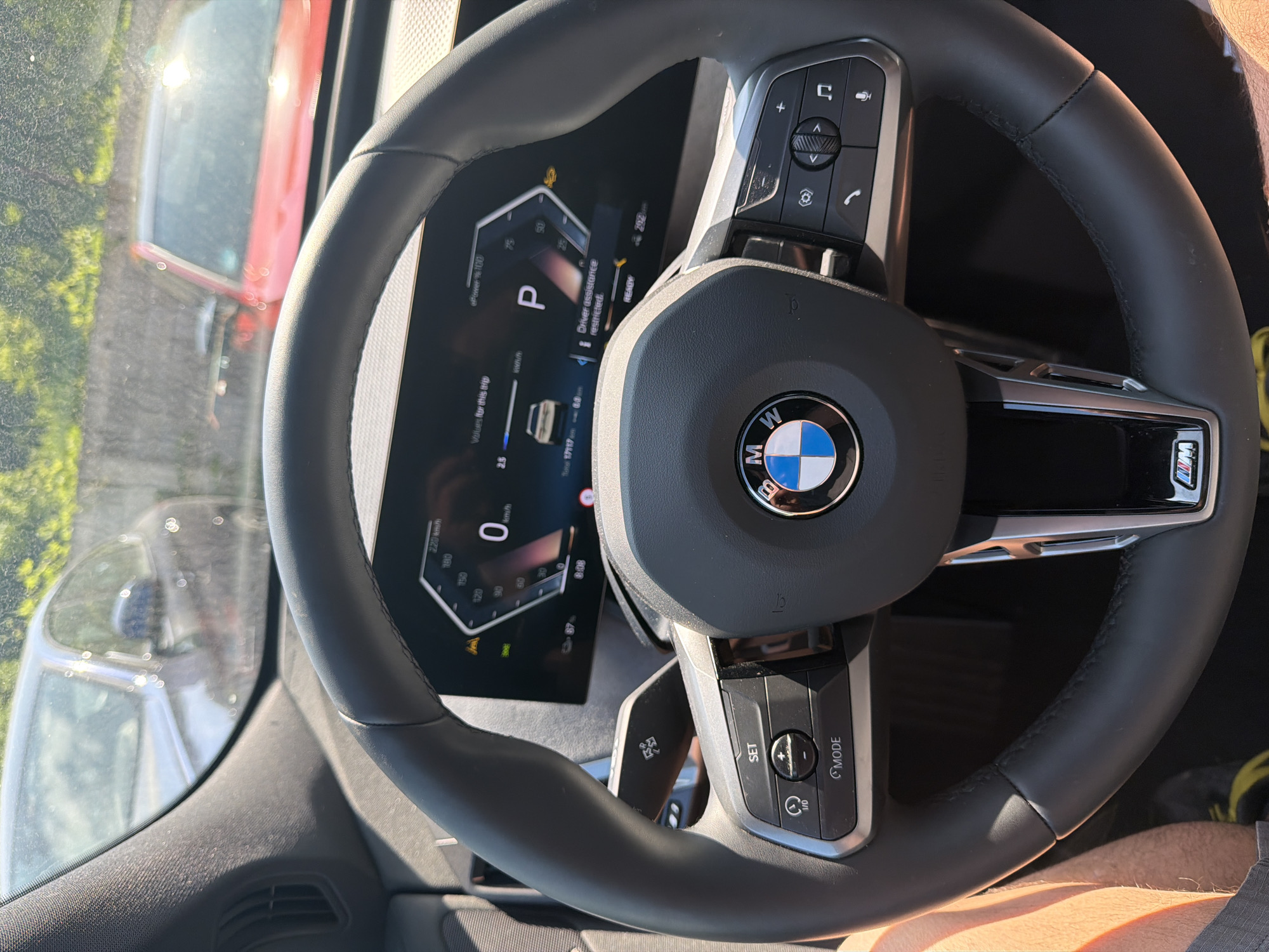 A first-person perspective shows the interior of a BMW, with the steering wheel and dashboard prominently in view. The dashboard display shows the car is in park ('P') and the driver assistance system is restricted. Outside the car, other vehicles and greenery are visible through the windshield, suggesting the car is stopped in traffic or parked. The photo appears to have been taken on a sunny day. No specific location in Burgas, Bulgaria is identifiable from the image.