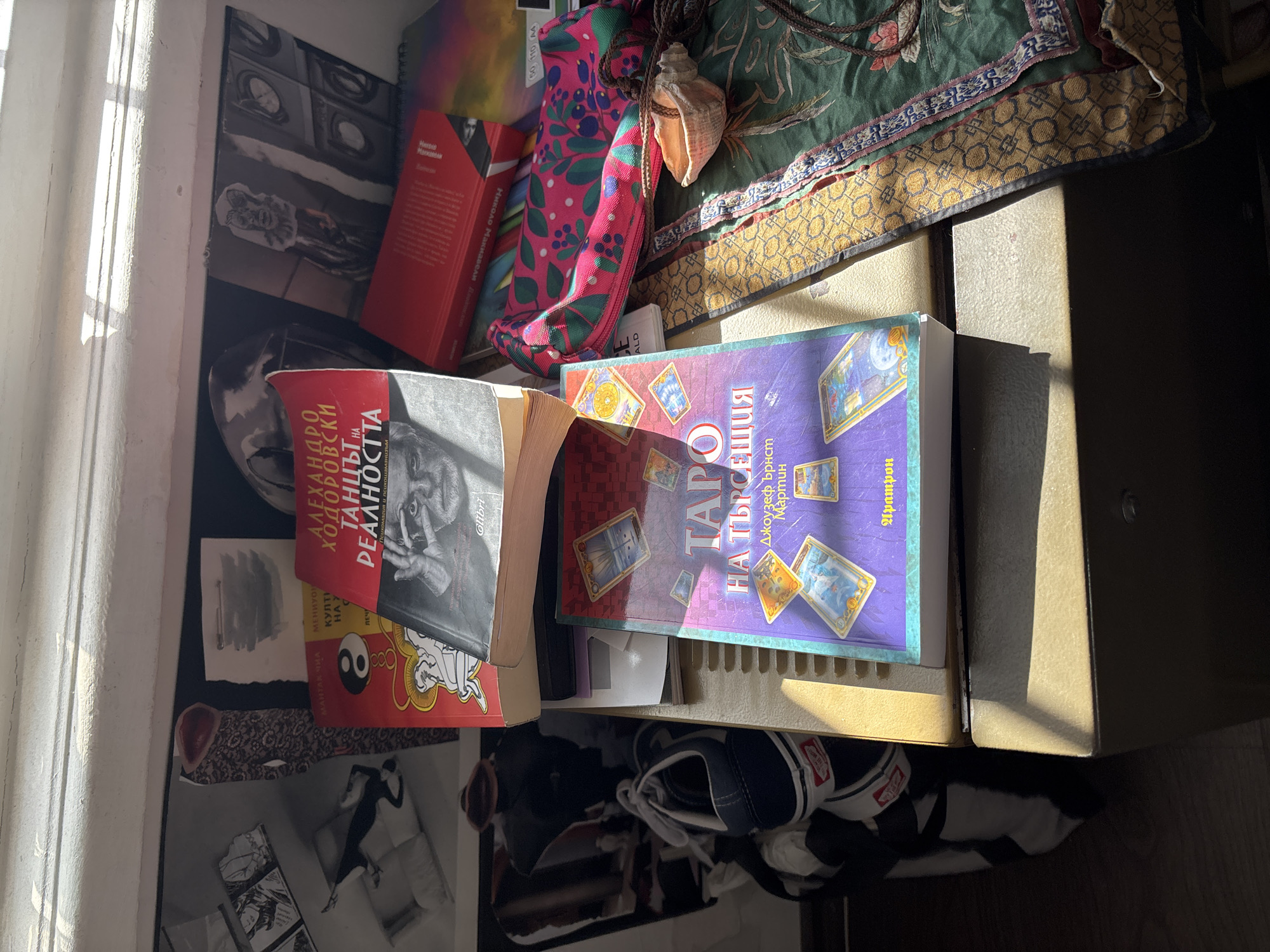 This media file captures an indoor scene featuring a cluttered surface, likely a desk or shelf, illuminated by strong, angled sunlight streaming from a window on the left. The image is rotated counter-clockwise, making the window appear on the left edge.
The prominent subjects are several books and personal items. Two large books are visible: one red, titled "ТАНЦЪТ НА РЕАЛНОСТТА" (The Dance of Reality) by "АЛЕХАНДРО ХОДОРОВСКИ" (Alejandro Jodorowsky), featuring a black and white image of a man's face. Another book has a purple and blue cover adorned with Tarot cards, titled "ТАРО НА ТЪРСЕЩИЯ" (Tarot for the Seeker) by "Джоузеф Ърнст Мартин" (Joseph Ernst Martin), published by "Аратрон." A smaller red book with "Николай Манол" (Nikolai Manol) is partially visible, as is a book by "МАНТАК ЧИА" (Mantak Chia).
Other objects include a vibrant pink and green patterned pouch resting on a green and gold patterned fabric. A seashell with a braided cord is also on the fabric. Below the books, a pair of dark blue and white Vans sneakers are partially visible. Various papers and prints are scattered, including one with a prominent Yin-Yang symbol and another with what appears to be manga art. A white window frame and curtain line the left side of the image.
No specific activity is taking place; the scene is a still life. The harsh shadows and direct sunlight suggest it is either morning or late afternoon, indicating sunny weather. The Cyrillic text on the books, specifically Bulgarian, aligns with the provided location context of Sofia, Bulgaria. The presence of books on psychomagic, tarot, and Taoist practices suggests an interest in esotericism and spirituality. The overall impression is one of a personal, lived-in space.