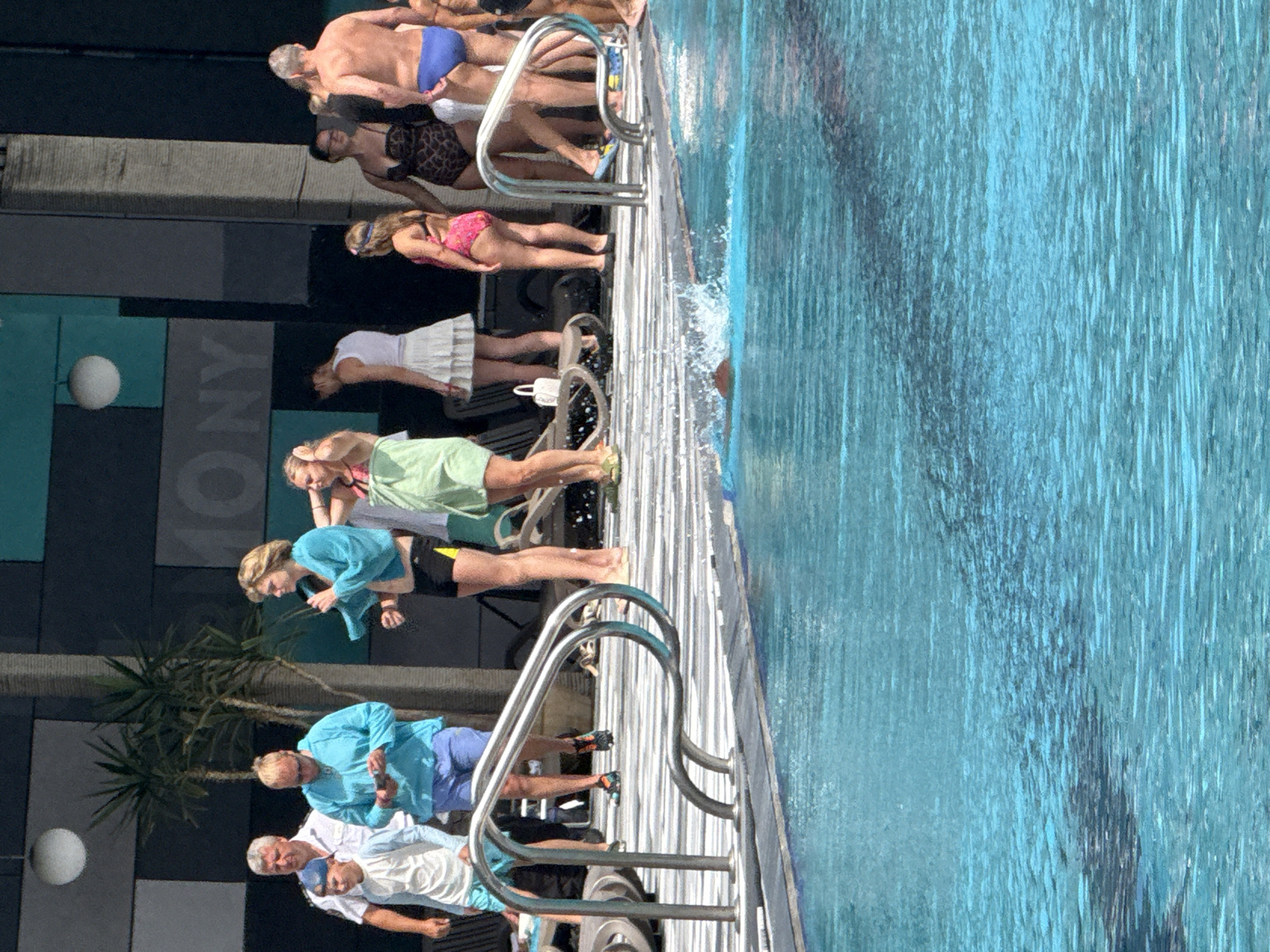 In the city of Массандровский поселковый совет, Ukraine, a group of people are gathered around a swimming pool on a sunny day. A man in a light blue long-sleeved shirt and blue shorts is holding a phone. To his left, a boy in a white t-shirt and blue shorts is standing. A man in a white shirt and black shorts is also visible. Another person, wearing a turquoise towel, is holding a microphone or similar object. A woman in a light green towel and a white skirt is standing next to them. A child in a pink swimsuit is also present. Further to the right, a woman in a leopard-print one-piece swimsuit and a man in blue swim trunks are standing. The swimming pool is filled with clear blue water, and some ripples are visible on the surface. A person is swimming in the pool, with their head submerged. The background consists of a building with a dark grey facade and teal accents.