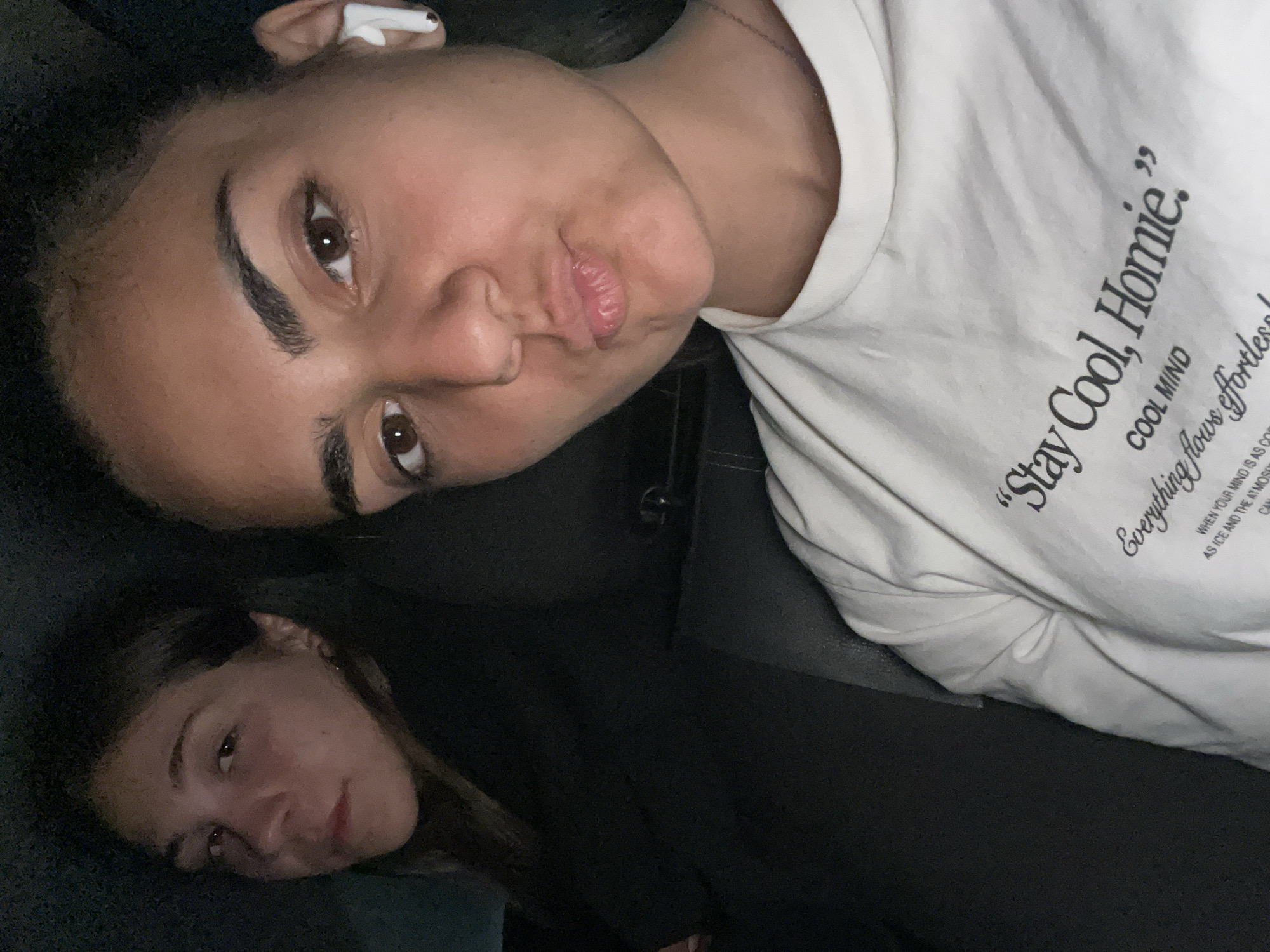 Two young women are in a vehicle, likely a car, based on the black interior. The woman closer to the top of the frame is wearing a white T-shirt with the words "Stay Cool, Homie. COOL MIND. Everything flows effortless". She is also wearing an earbud. The second woman is wearing a dark, possibly black, garment. No information is available about the location of this image in Debelt, Bulgaria.