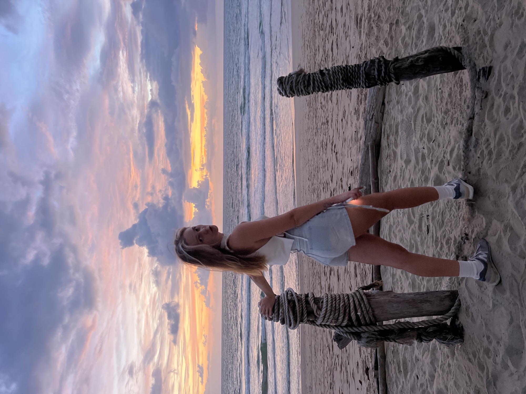 A young Caucasian woman stands on a sandy beach in Choeng Thale, Thailand, during sunset. She is facing away from the camera, looking over her left shoulder towards the viewer. She has long, blonde hair and is wearing a white halter top and light blue denim shorts. She has white socks and gray sneakers on. She is leaning on a wooden post wrapped with thick rope, which is part of a rustic structure on the beach. The ocean is behind her, with gentle waves breaking on the shore. The sky is filled with dramatic clouds illuminated by the setting sun, casting hues of orange, pink, and purple across the scene. The overall atmosphere is serene and picturesque, characteristic of a tropical beach at dusk.