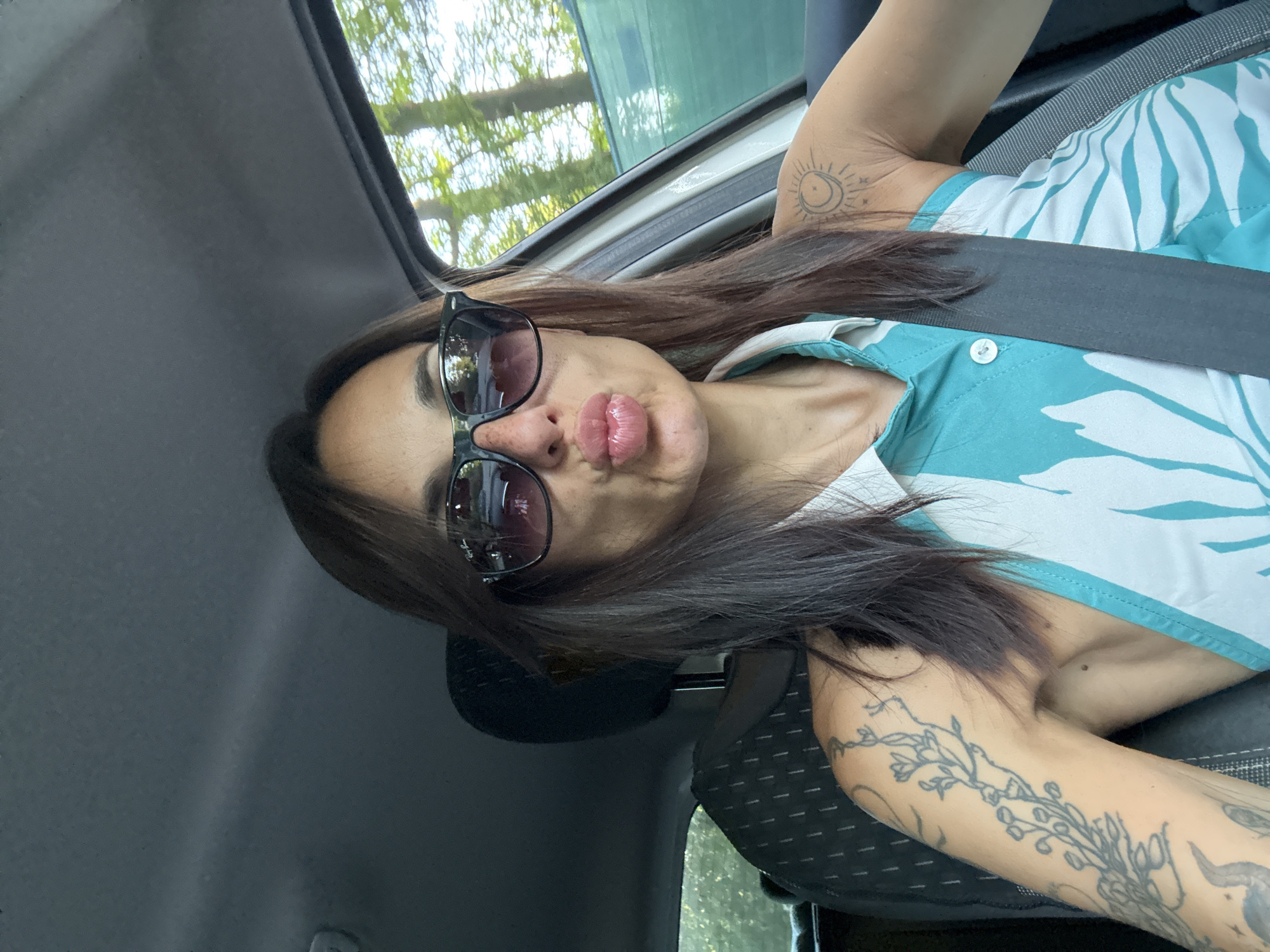 A woman in sunglasses and a blue and white patterned top is sitting in the driver's seat of a car, making a kissy face at the camera. She has several tattoos on her arms, including a floral design on her left arm and a celestial design on her right shoulder. She is wearing a seatbelt. The interior of the car is visible, as is the scenery outside the window, which appears to be trees and foliage, suggesting an outdoor setting. The scene is likely in a residential area or along a tree-lined road, possibly in or around the city of Pleven, Bulgaria.