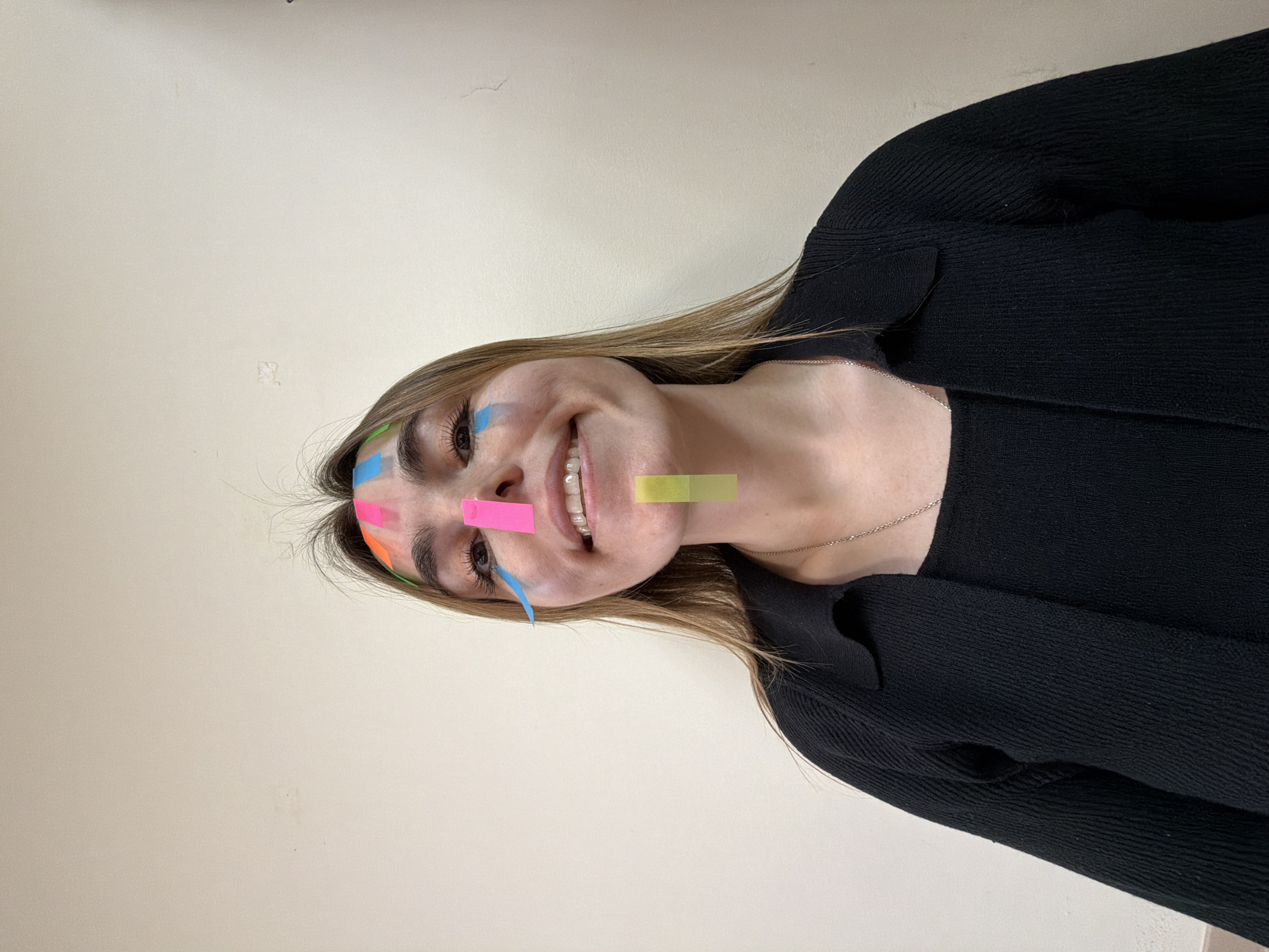 A young woman smiles at the camera. She has light brown hair and is wearing a black sweater. Various colored sticky notes are adhered to her face. The background is a plain, off-white wall. The image does not appear to be taken in Izhevsk, Russia.