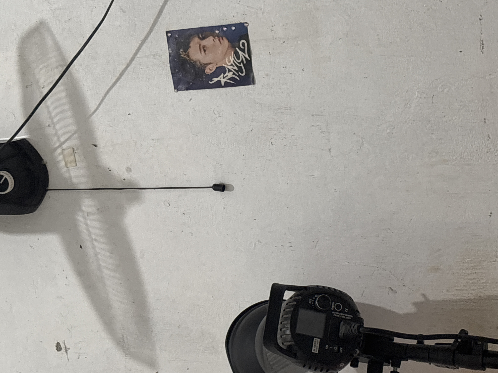 The image depicts a close-up view of a rough, off-white textured wall in what appears to be an indoor setting. Dominating the lower left of the frame is a black photography light on a stand, with its control panel visible. The light is connected to a power cord. To the right, a small poster featuring a portrait of a young man with dark hair and a white graphic overlay is pinned to the wall. Above and to the left of the poster, a fan is mounted to the ceiling, and its shadow is cast prominently across the wall. A dark cord hangs from the fan, with a black cylindrical object at its end. The overall lighting suggests an indoor environment, likely a studio or a room with minimal natural light. The presence of photography equipment indicates a potential use for creative purposes. The rough texture of the wall and the general appearance of the space suggest it might be a utilitarian or unfinished area. The location of North Jakarta, Indonesia, is provided as context, implying a specific geographical setting for this scene.