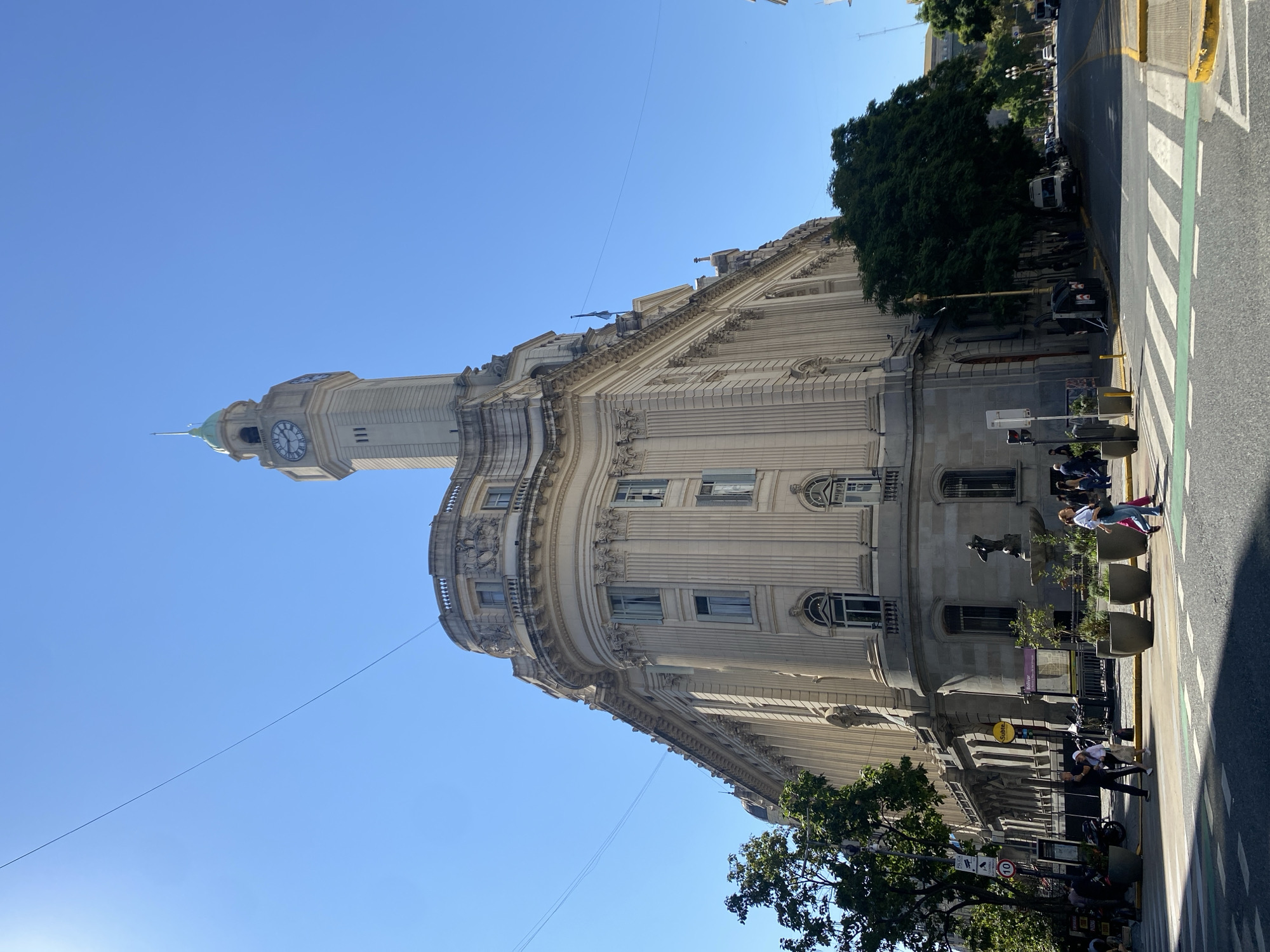The image shows the Palacio de Correos, located in Buenos Aires, Argentina. It is a large, ornate building with a clock tower. The building occupies an entire block. It is daytime, with clear blue skies. People are walking on the sidewalk in front of the building. Cars are parked along the street. Trees line the sidewalk. The photo is taken from a low angle.