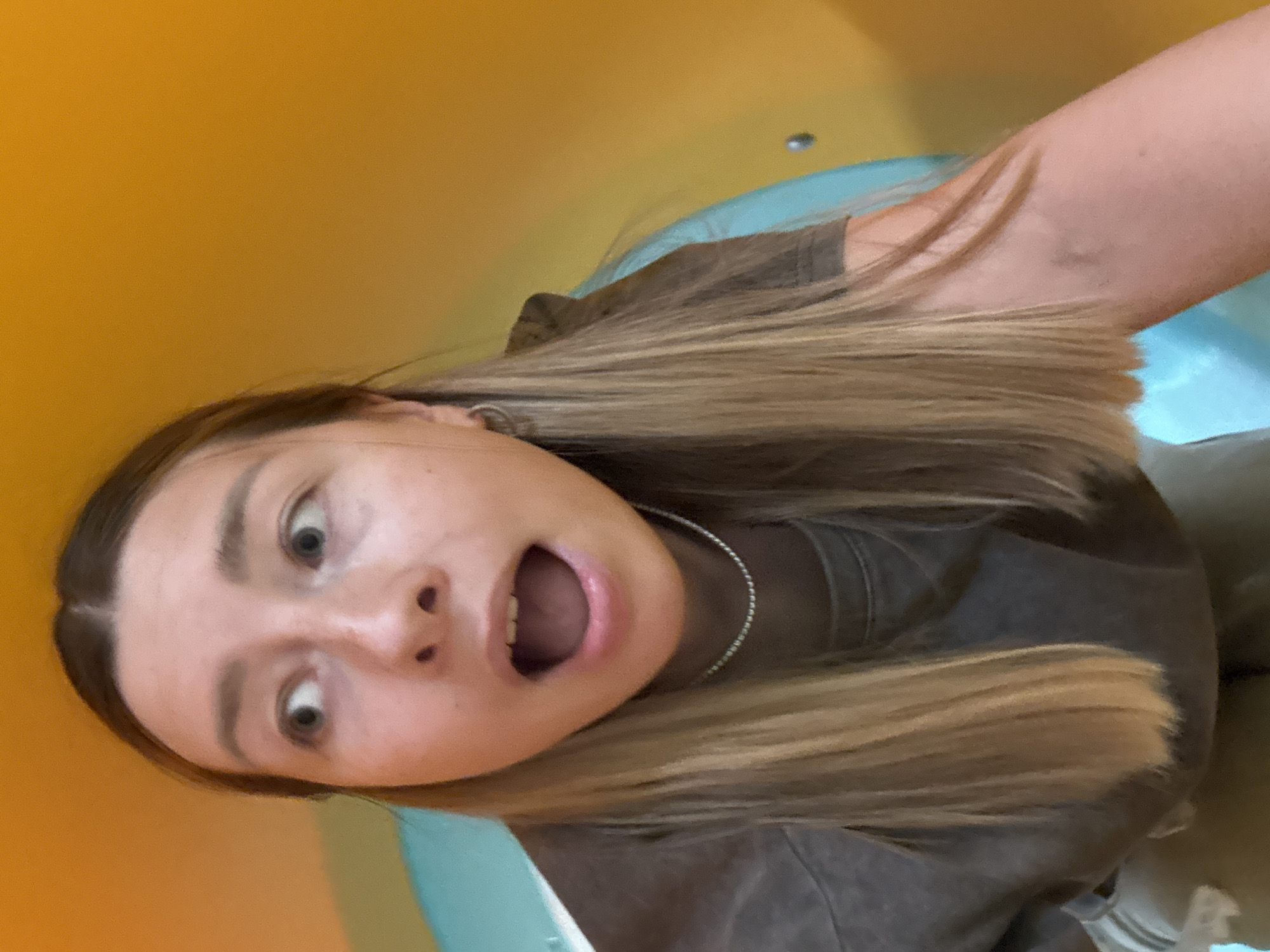 A woman with shoulder-length blonde hair is captured in a close-up, angled selfie. She is wearing a dark-colored t-shirt and a silver chain necklace. Her mouth is open in an expression of surprise or shock, and her eyes are wide. The background consists of a blue and yellow surface, possibly a slide or play structure. The location is not identifiable as Khimki, Russia from the image alone.