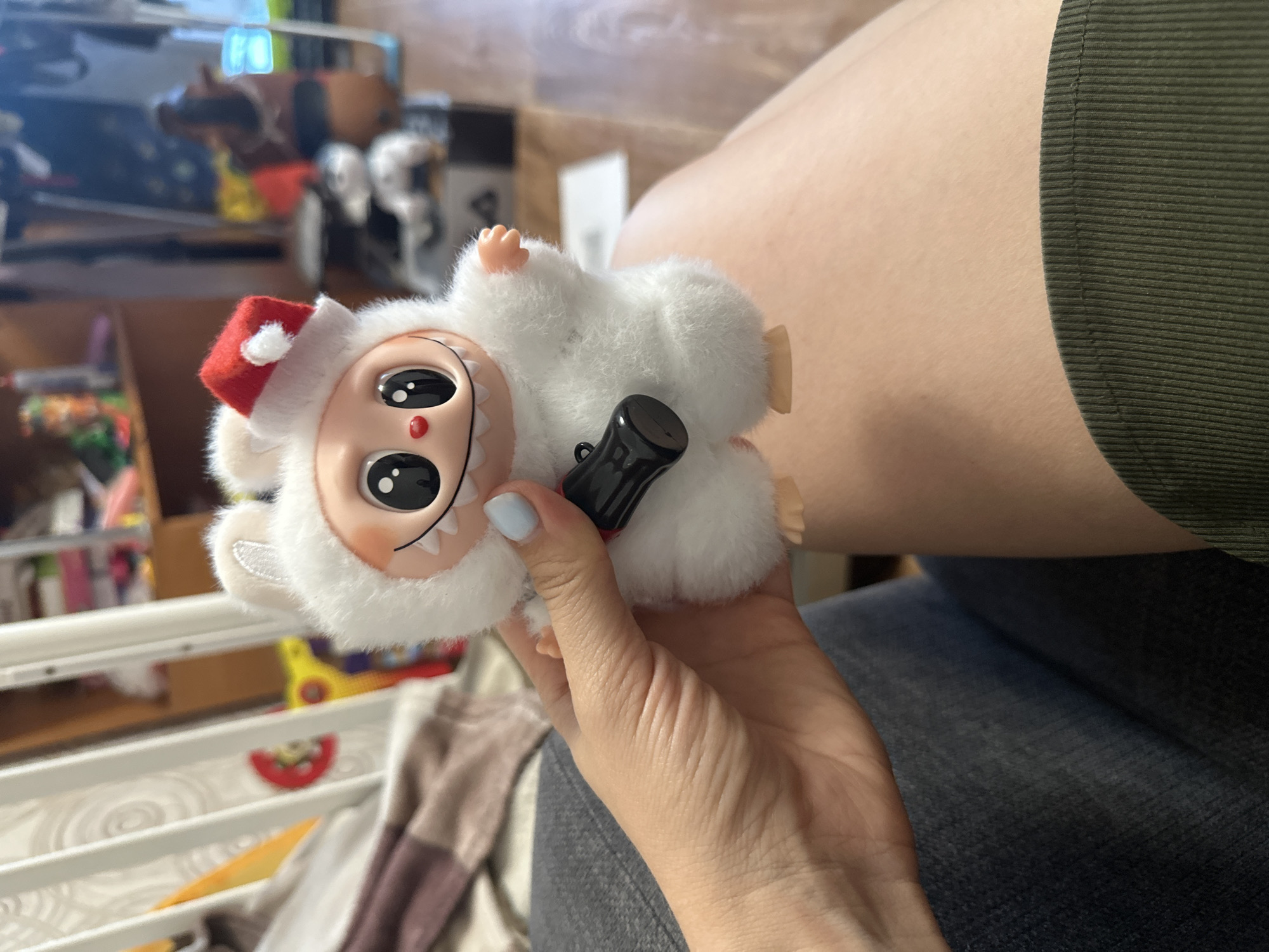 A person holds a plush toy with a white fluffy body, large black eyes, a red and white Santa hat, and a small black accessory attached to its chest. The person's hand, with pale blue nail polish, is visible supporting the toy. The background shows a cluttered indoor environment with shelves of items and furniture, suggesting a domestic setting. The overall lighting indicates an indoor scene with some ambient light.