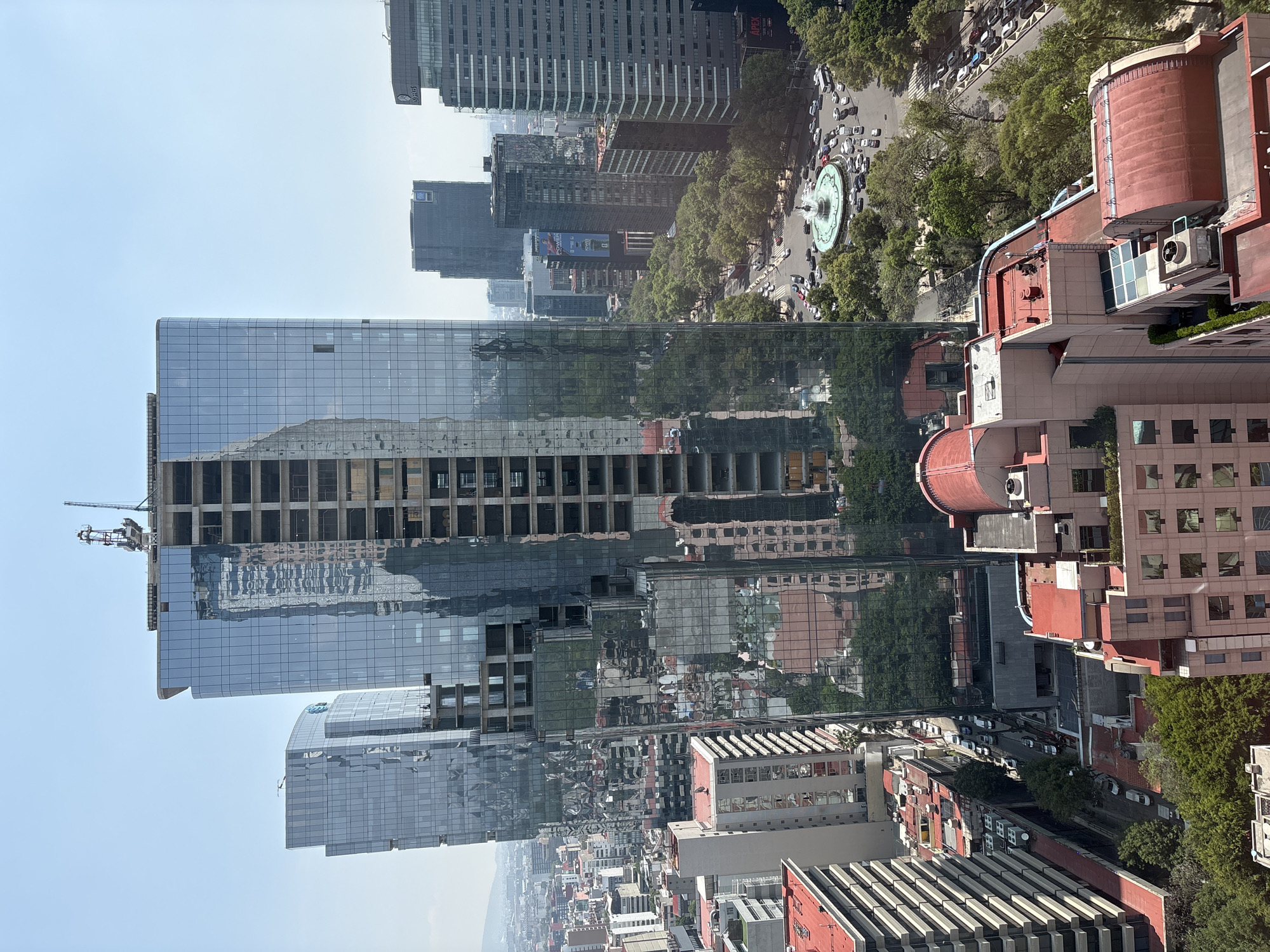 The image captures a high-angle view of a bustling urban landscape in Mexico City, bathed in bright daylight under a clear blue sky. A prominent, modern glass skyscraper, still under construction with visible concrete framework and a crane atop, dominates the left side of the frame. Its reflective facade mirrors the surrounding city, including other buildings, trees, and the sky.
In the mid-ground, a circular fountain with spraying water is a focal point on a busy street, surrounded by moving cars and lush green trees. Several other dark-colored skyscrapers are visible in the background, with discernible text "MEX" and "APEX" on some of their facades. To the right and in the foreground, a cluster of buildings with distinct red-tiled or flat red roofs, some featuring air conditioning units and rooftop equipment, adds architectural variety. The cityscape extends into a hazy distance, suggesting a vast metropolitan area. The active traffic around the fountain and the ongoing construction on the skyscraper indicate a dynamic and growing urban environment. The time of day appears to be late morning or early afternoon, given the bright illumination.