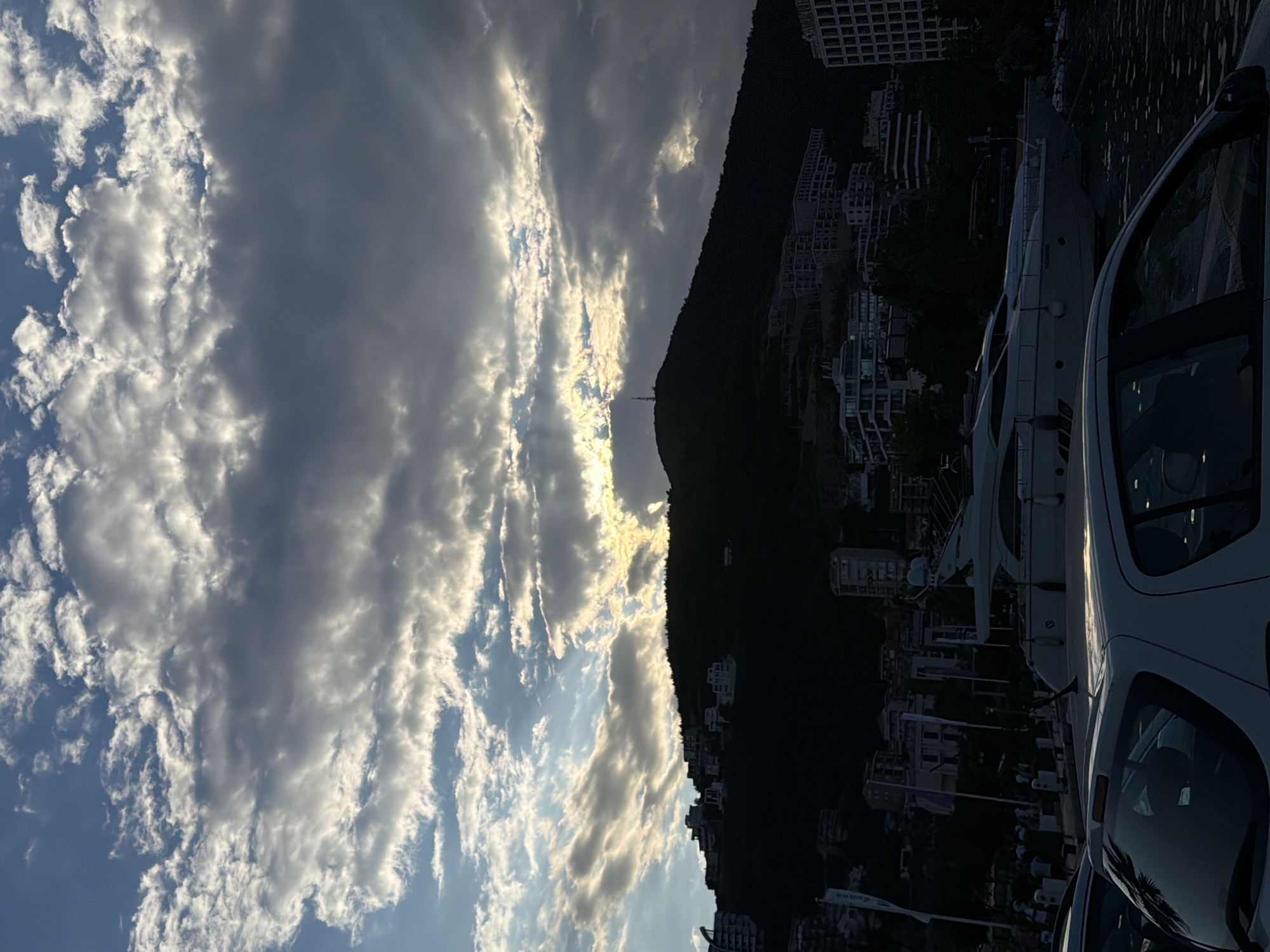 The image depicts a waterfront scene in Budva, Montenegro, likely taken at dusk or dawn. The sky is filled with dramatic clouds, with patches of sunlight breaking through, creating a luminous effect. A dark, silhouetted mountain or hillside dominates the right side of the frame, dotted with numerous buildings that are part of a coastal town or resort.
In the foreground, a white vehicle is partially visible, suggesting it is parked by the waterfront. Behind the vehicle and along the coastline, several white yachts are moored. The overall impression is one of a serene yet vibrant coastal environment at the transitional hours of the day, with the natural beauty of the sky and landscape contrasting with the man-made structures and vessels. The lighting suggests either sunrise or sunset, casting long shadows and highlighting the textures of the clouds. There are no discernible people or text in the image.