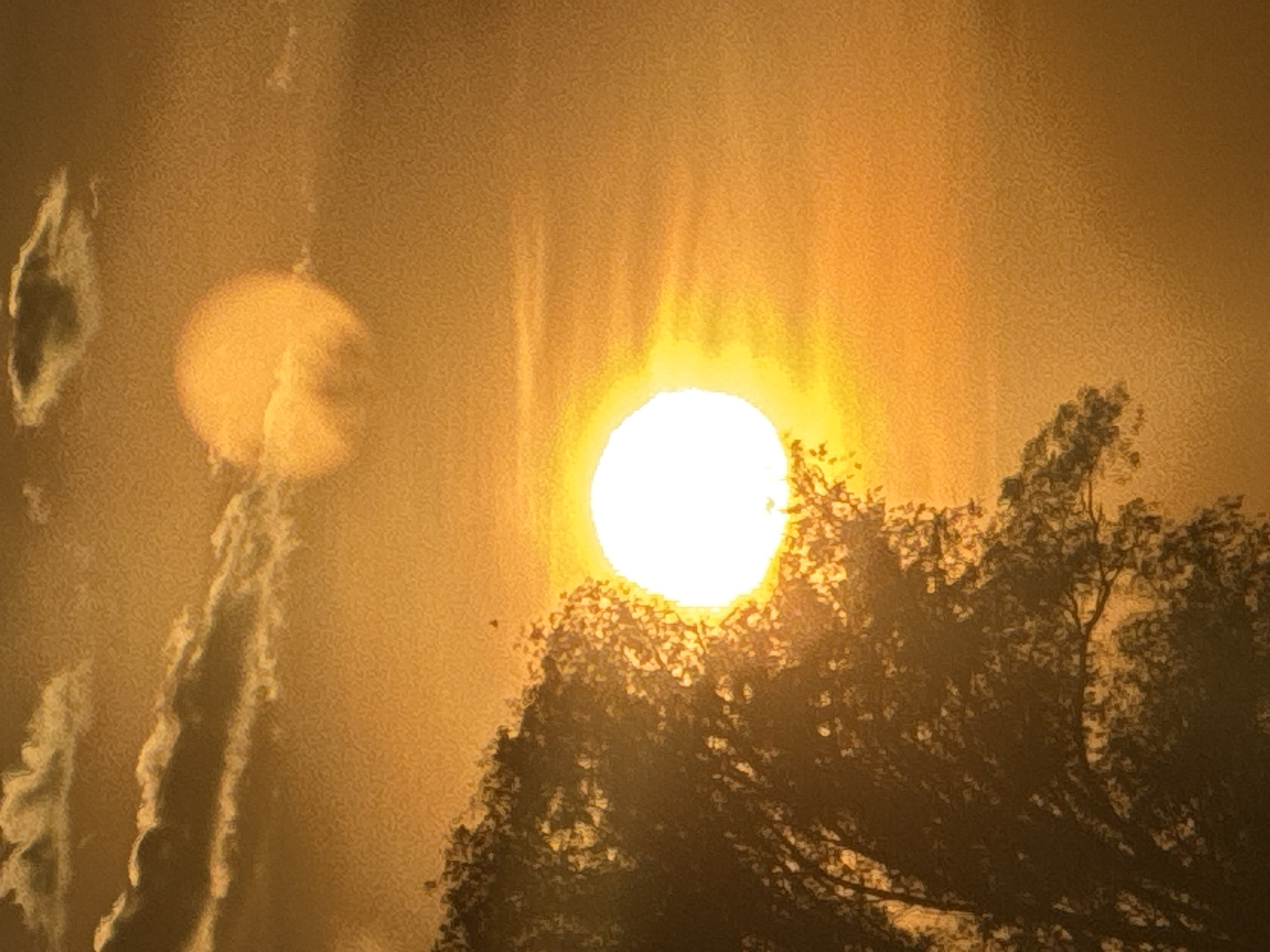 The image, taken in Ohuivka, Ukraine, displays an orange and yellow sky during what appears to be either sunrise or sunset. A brilliant, circular sun is prominently positioned in the lower-left quadrant, partially obscured by the dense, dark silhouette of a large tree occupying the left side of the frame. Above and to the right of the primary sun, a larger, diffused orange circular glow is visible, consistent with an optical lens flare or reflection. The upper-left portion of the sky contains several dark, elongated clouds with bright edges. Faint, horizontal atmospheric bands or wispy clouds are present across the middle-right of the frame. No human or animal subjects are visible. The entire image exhibits a pronounced grainy texture.