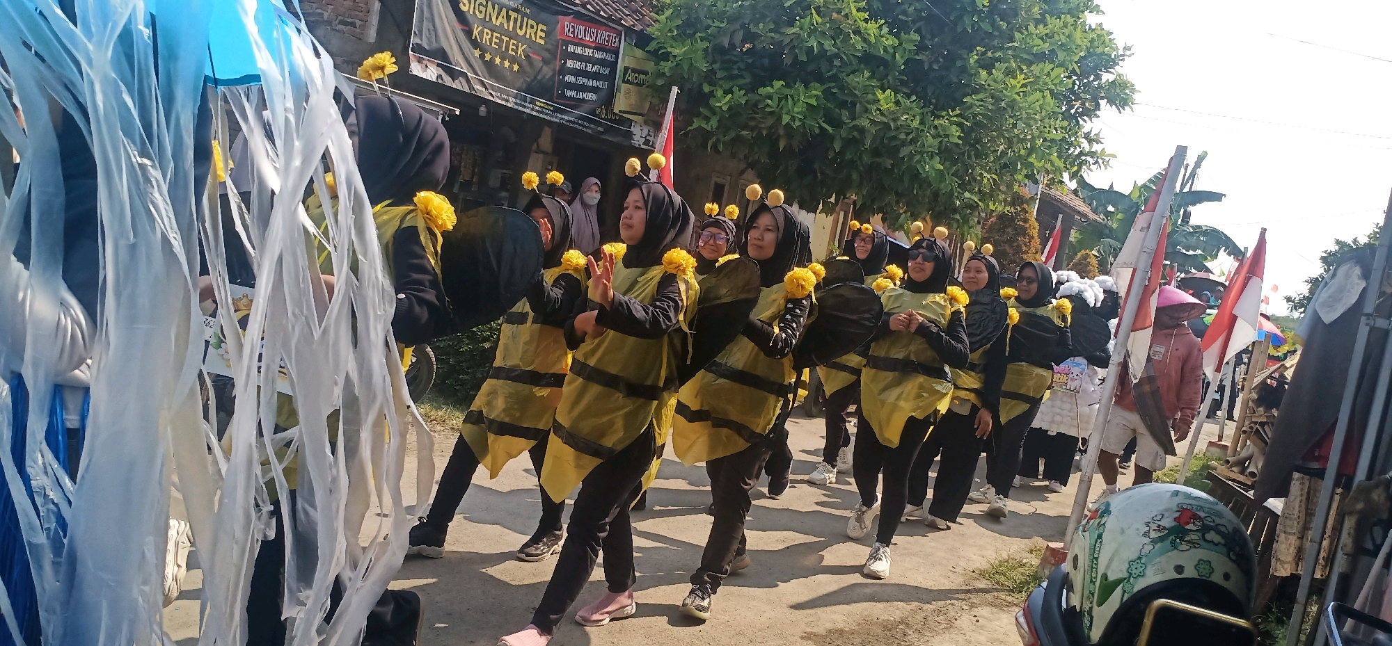 A group of people dressed as bees are participating in a parade in Kincang, Indonesia. The individuals are wearing black clothing with yellow bee-striped vests and yellow pom-pom antennae. Some are carrying yellow pom-poms in their hands. The parade appears to be on a street lined with trees and buildings. Indonesian flags are visible in the background. One person in the foreground is holding a blue and white decoration that resembles jellyfish tentacles. Another person is wearing a pink conical hat and a pink hooded jacket. The overall scene suggests a festive community event.