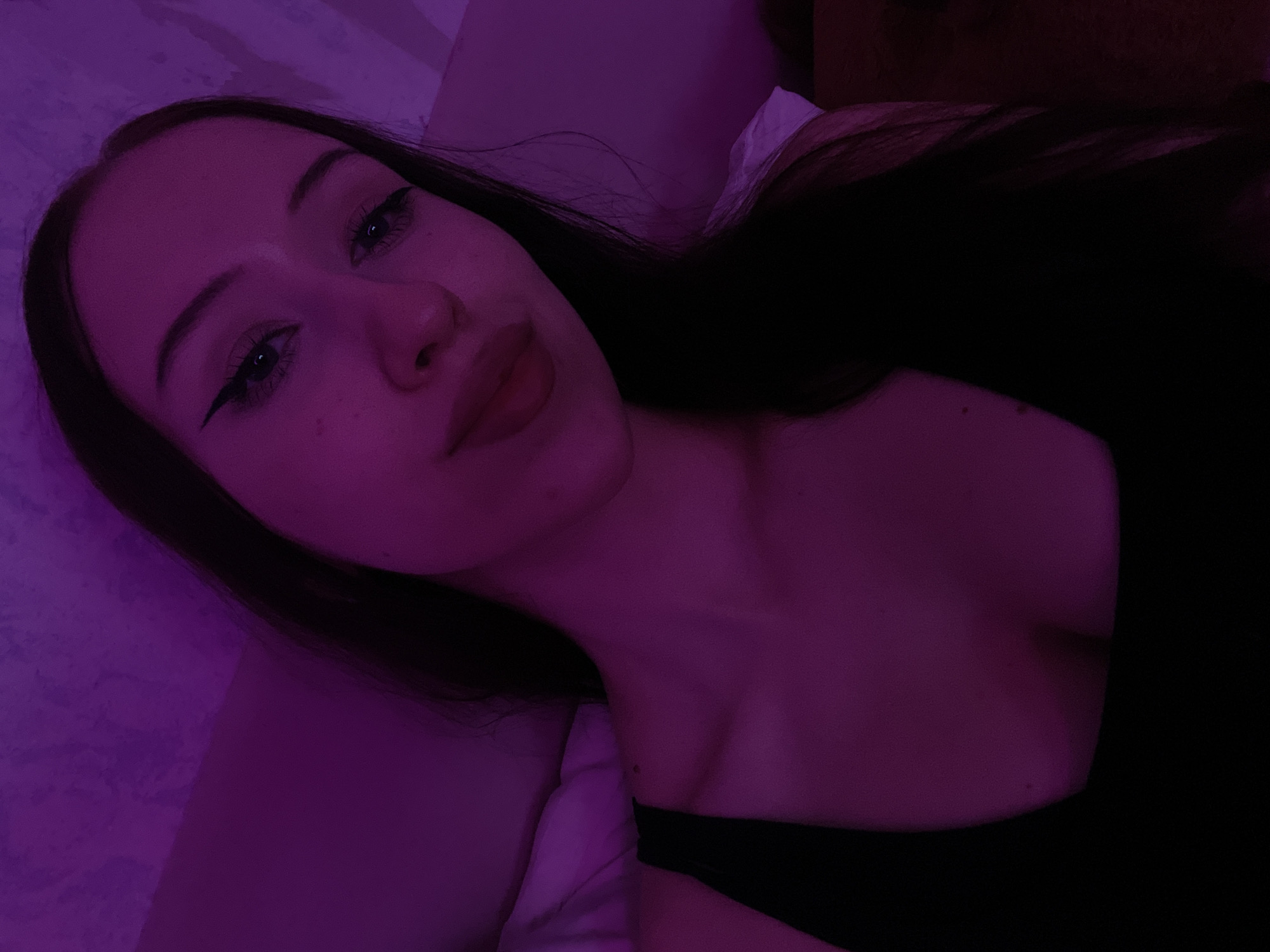 A young woman with long dark hair and winged eyeliner is captured in a close-up selfie. She is wearing a black top and is illuminated by a purple light. The background appears to be a textured wall. The image does not provide any information about the location in Votkinsk, Russia.