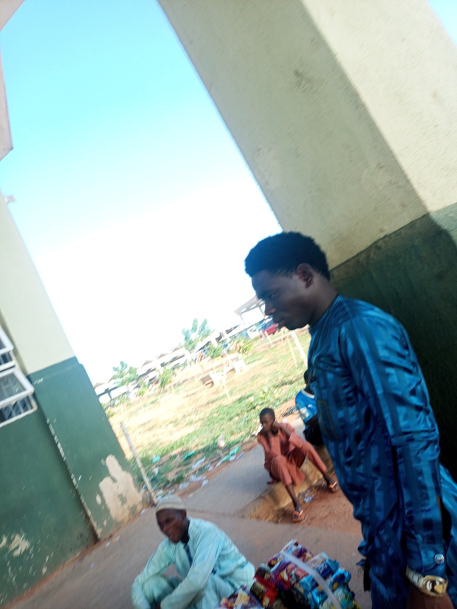 Three individuals are visible outdoors during daylight. A person in a blue, shiny tunic is partially in view, standing to the right. In the middle distance, another individual is seated, wearing a reddish-brown garment and sandals. In the foreground, another person sits on the ground wearing a light blue outfit. Next to them is a pile of assorted goods, possibly for sale. The background contains a grassy field and distant buildings under a bright sky. The image appears to have been taken at a slightly skewed angle.