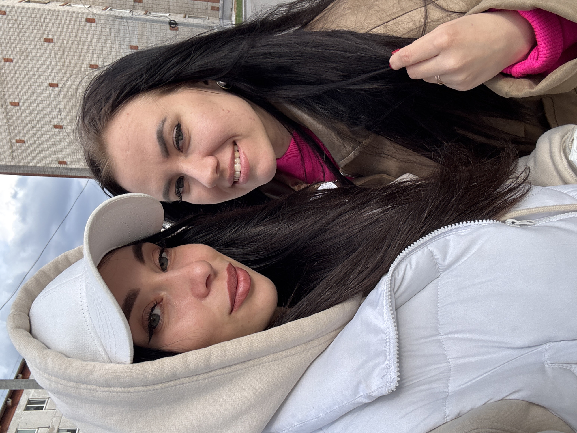 This close-up outdoor photo, set in Chaikovsky, Russia, features two young women posing together on what appears to be an overcast day.
The woman positioned in the upper right part of the frame has long, dark hair. She is smiling broadly, revealing her teeth, and wears a bright pink top under a light brown or tan jacket. Her left hand, adorned with red nail polish and a ring on her ring finger, gently holds a strand of her dark hair.
The second woman, positioned in the lower left, also has long, dark hair and looks towards the camera with a more neutral, composed expression. She is wearing a white baseball cap, a light beige hooded sweatshirt, and a white puffer jacket with a visible zipper down the front.
The background shows a large building with light-colored, possibly brick or concrete, textured walls and a few small, dark windows. Above the building, the sky is predominantly cloudy. The overall mood is casual and friendly. There is no visible text in the image.
