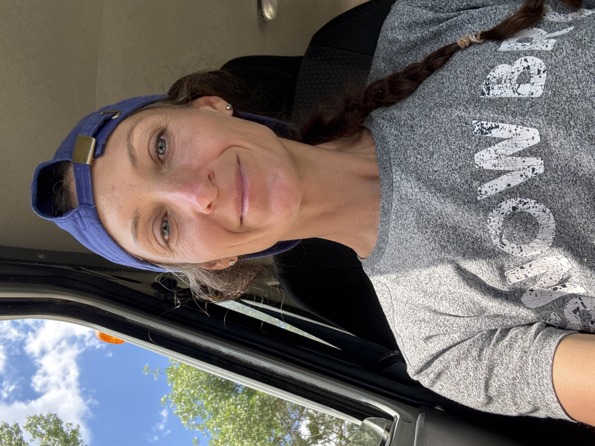 A woman with brown hair in a braid, wearing a grey t-shirt and a blue baseball cap, is pictured from the chest up. She is looking at the camera and smiling. The background shows the interior of a vehicle, with a glimpse of trees and the sky through the window. There are no visible elements that would indicate the location as Pushkino, Ukraine.