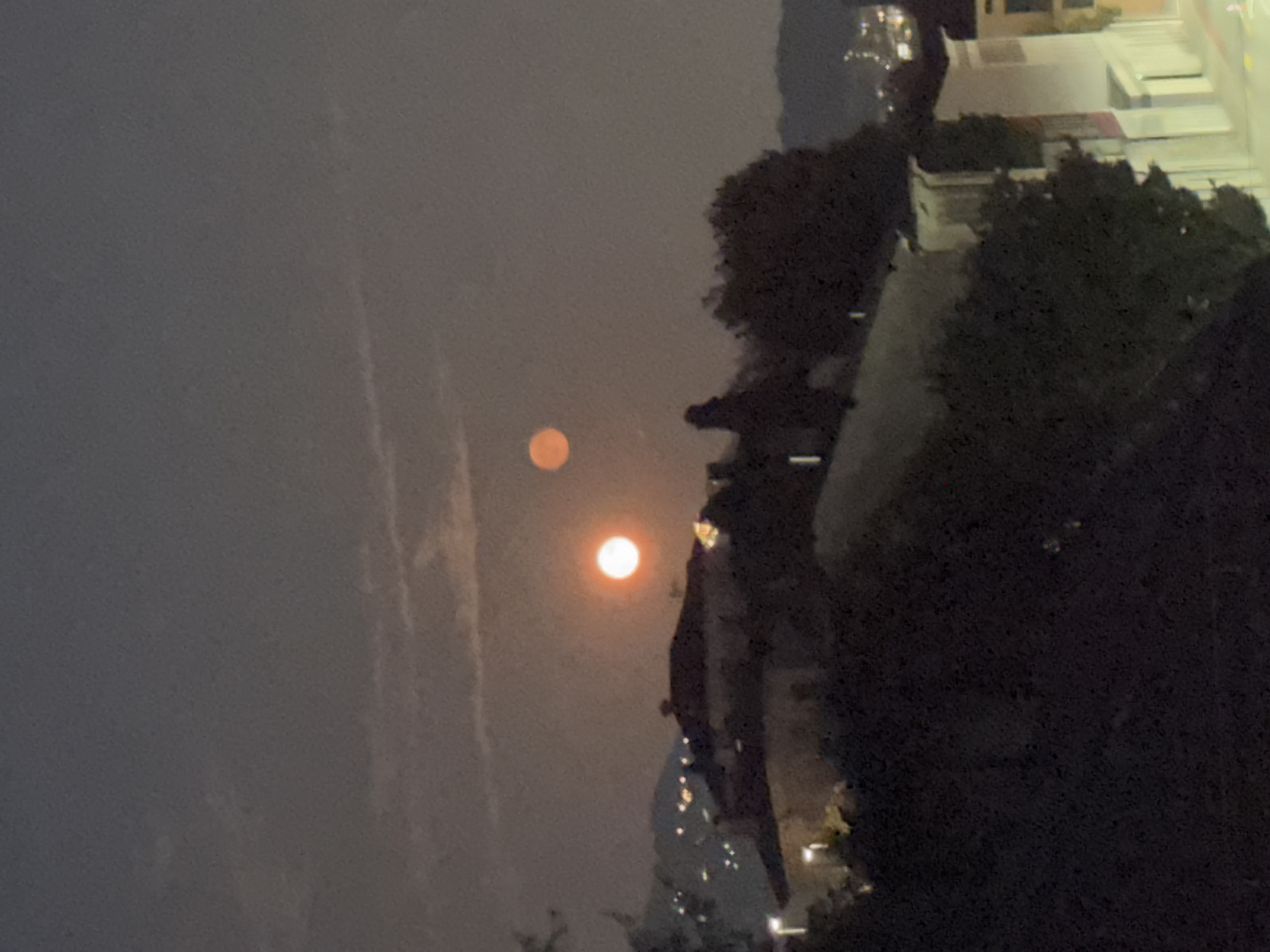The image captures a nighttime scene in Balerna, Switzerland, with a dark sky and a faint haze. Two celestial bodies, appearing as full moons, are visible in the sky. One moon, a bright orange orb, is positioned higher and to the right of the other. The second moon, slightly dimmer and with a reddish hue, is located below and to the left of the first.
In the foreground, silhouetted against the dimly lit sky, are the rooftops of buildings and a dense cluster of trees. The architecture suggests a European urban or semi-urban setting. Distant lights from what appear to be streetlights or windows twinkle on a hillside in the mid-ground, indicating the presence of a town or settlement. The overall atmosphere is serene and slightly ethereal due to the prominent lunar presence. There are no people or discernible animals visible in the frame.