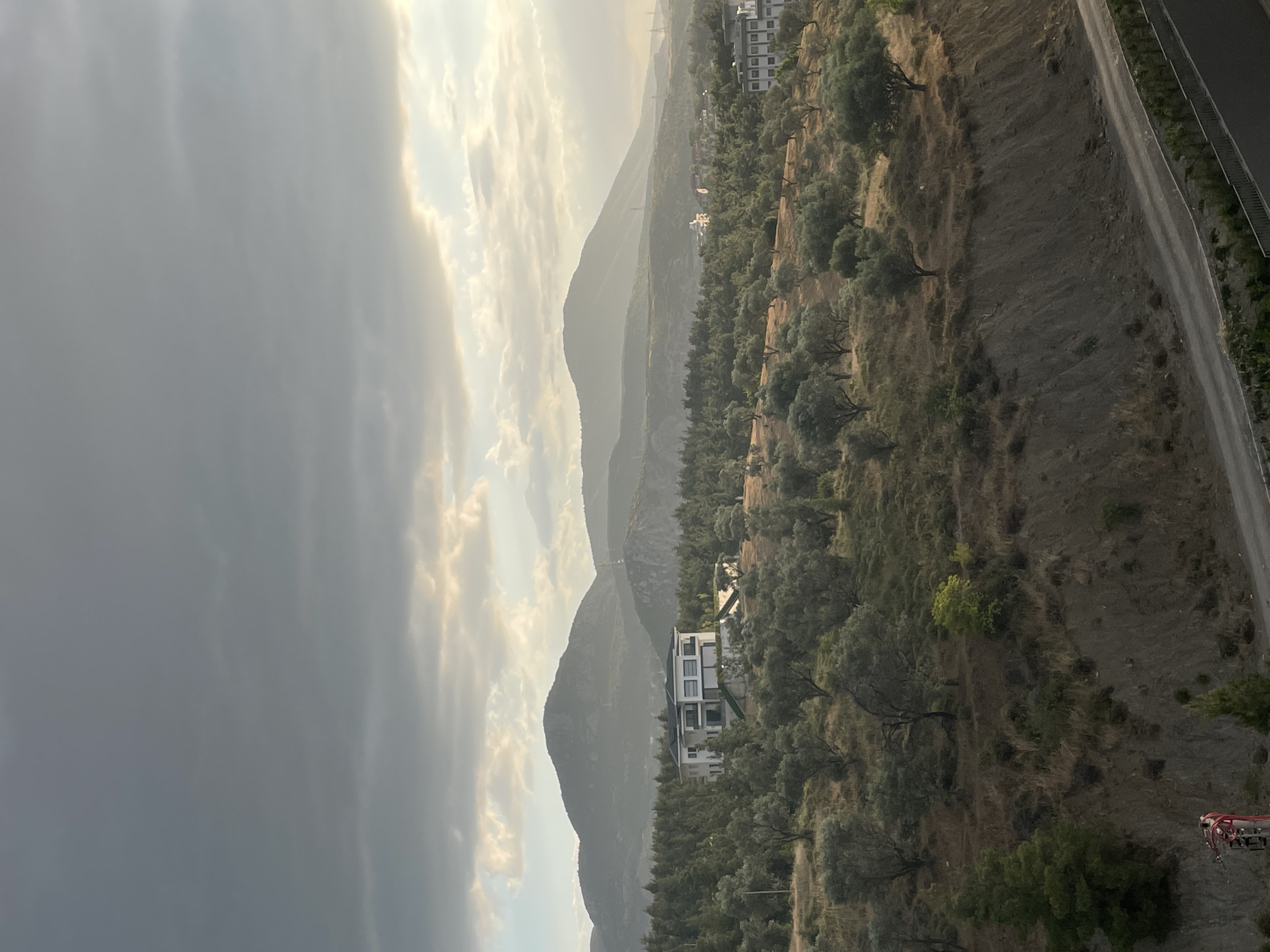 The image depicts a panoramic view of a hilly landscape with a prominent modern house nestled amongst olive trees. The scene is bathed in the soft, diffused light of what appears to be late afternoon or early morning, with dramatic, cloudy skies overhead. The foreground features a steep, eroded slope with dry vegetation and a paved road winding along its base. Further back, an orchard of olive trees dominates the mid-ground, with their distinctive gnarled trunks and silvery-green foliage.
A striking contemporary villa, characterized by its white facade, dark-framed windows, and flat rooflines, stands out in the center of the frame, surrounded by the olive grove. In the distance, layered mountain ranges recede into a hazy horizon, their forms softened by atmospheric perspective. On the far right, a multi-story building with a light-colored facade and numerous windows suggests a residential or institutional complex. The overall impression is one of rural tranquility, punctuated by modern architectural elements.
While the specific location within Yunus Emre, Turkey is not explicitly identifiable from visual cues alone, the scene is consistent with the hilly terrain and agricultural landscapes found in the region, particularly areas with olive cultivation. There are no people or discernible human actions captured in the image. The environment is natural and serene, with a clear emphasis on the interplay between the built and natural elements. The clouds in the sky suggest potential for changing weather, adding a dynamic quality to the scene.