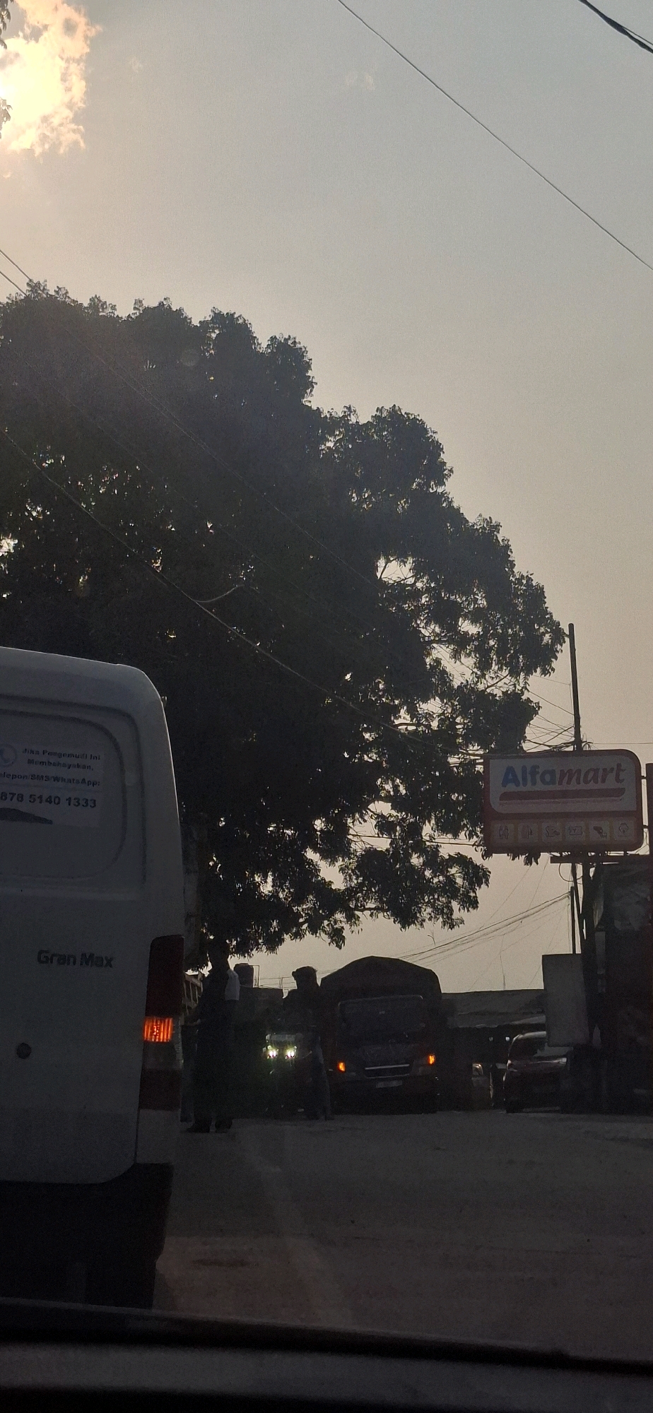 This image captures a street scene in Sirnagalih, Indonesia, at what appears to be dusk or dawn due to the soft, hazy light and the warm glow of the sky. The foreground is dominated by the back of a white van, identified by the text "Gran Max" on its side. On the van's rear window, there is text that reads: "Jika Pengemudi ini Membahayakan, Telepon/SMS/WhatsApp: 878 5140 1333".
Beyond the van, traffic is visible, including a dark-colored truck with its headlights on and another vehicle further back. Several silhouetted figures are present, some standing and others possibly near or inside the vehicles. Overhead, a large, dense tree with dark foliage casts shadows, and power lines crisscross the sky.
To the right, a prominent sign for "Alfamart" is clearly visible, indicating a commercial presence in the area. The overall atmosphere is one of a typical Indonesian street scene, possibly during a period of congestion or waiting. The muted colors and the soft light suggest a calm, perhaps slightly melancholic mood. The presence of the Alfamart sign provides a concrete geographical clue, suggesting this is a public area with access to convenience stores.