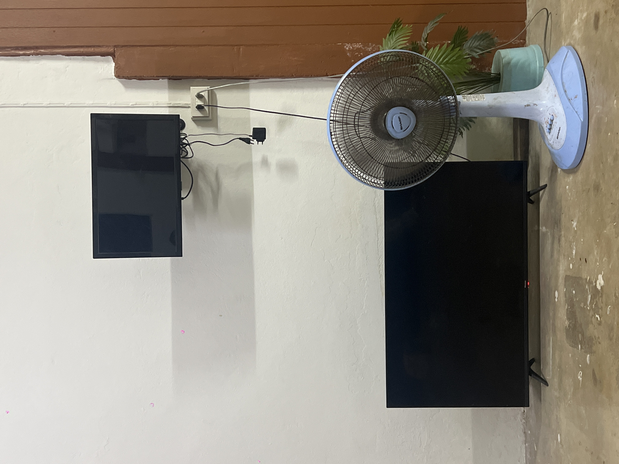 The image depicts an indoor scene, possibly a room in Thung Tako, Thailand. A white wall serves as the backdrop for two black flat-screen televisions. One television is mounted high on the wall, while the other rests on the floor. A blue and white electric fan, branded "SHARP," stands to the right of the lower television. The fan has a circular grille and a stand. Behind the fan, a green potted plant is visible, with its leaves extending upwards. Electrical outlets and cords are present on the wall near the upper television, with a power adapter hanging below. The floor appears to be concrete with some scattered white debris. A section of a brown wooden doorframe is visible on the far right edge of the image.