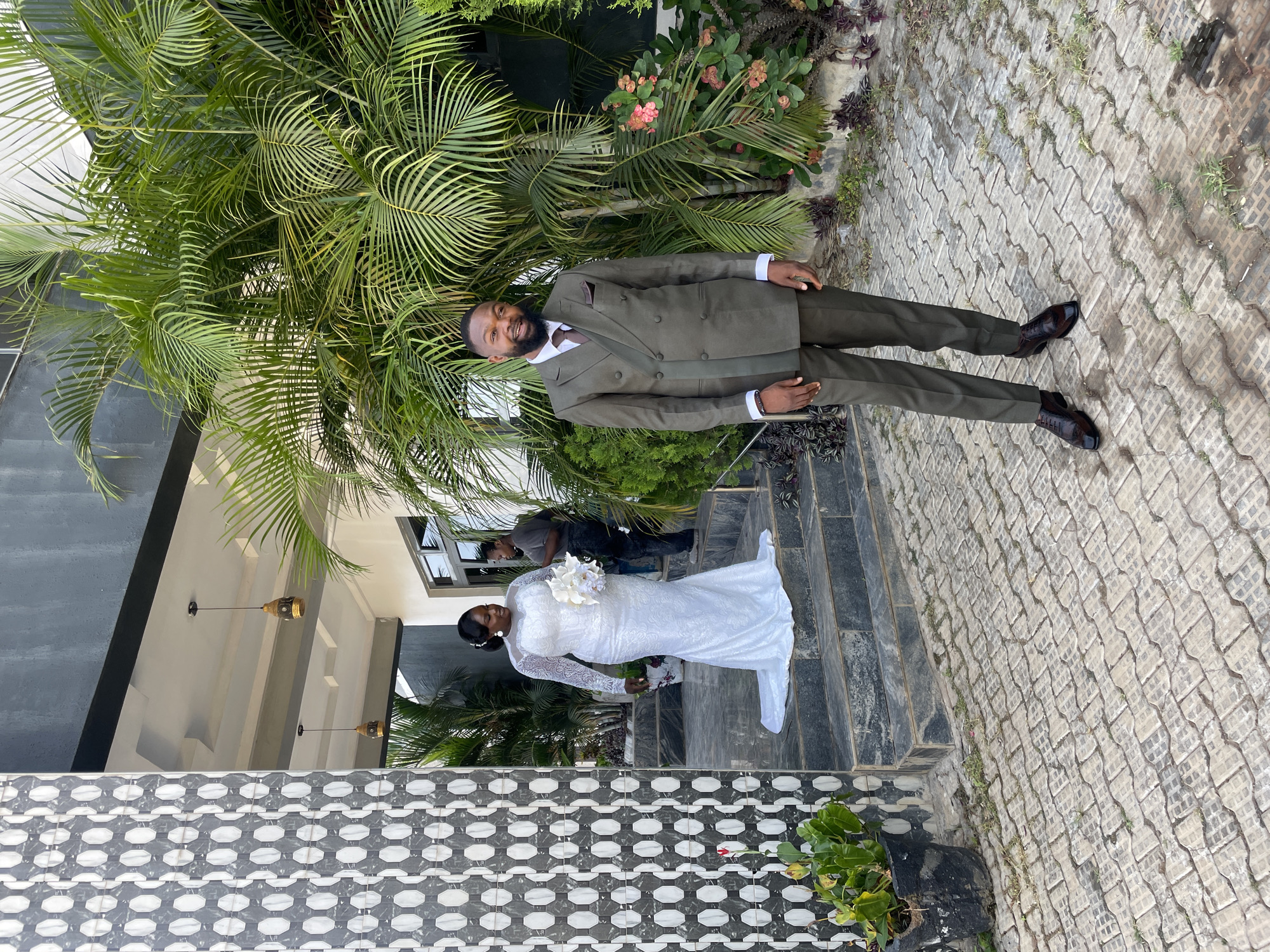 A wedding is being photographed outdoors in Jos, Nigeria. A man in a green suit stands next to a palm tree, while a woman in a white wedding gown stands on stairs leading from a building with patterned tiling. She holds a white bouquet. A photographer is present in the background, focusing on the bride. The surrounding environment consists of buildings, foliage, and paved ground.