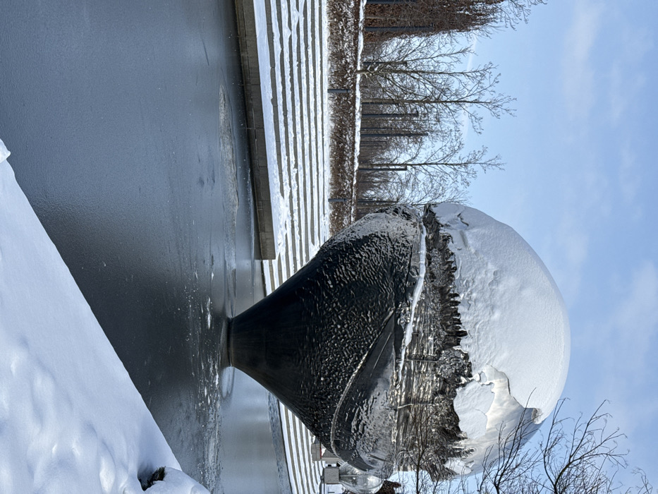 The image depicts a winter scene in the city of Plodorodny, Russia, on a clear day under a light blue sky. A large, metallic, spherical sculpture, textured and reflective, serves as the central subject. The upper portion of the sculpture is heavily blanketed with white snow, showing uneven edges and formations suggestive of melting or refreezing. Its lower half is dark and textured, tapering downwards into a conical base. This sculpture is situated within a body of water, which is predominantly frozen with a smooth, dark sheet of ice. Immediately surrounding the sculpture's base, the ice appears slightly disturbed or melted.
To the left and rear of the sculpture, a series of wide, shallow, snow-covered stone or concrete steps ascend from the water's edge. Beyond these steps, a landscaped area is visible, featuring numerous bare deciduous trees and some darker, possibly evergreen, taller trees or shrubs. A low, snow-covered hedge or embankment runs horizontally along the base of this tree line. Several dark, slender vertical posts are spaced throughout the background landscaping. In the immediate foreground, to the right, a bank of deep white snow slopes diagonally upwards from the frozen water. No individuals are present in the scene, and no overt actions or interactions are depicted.