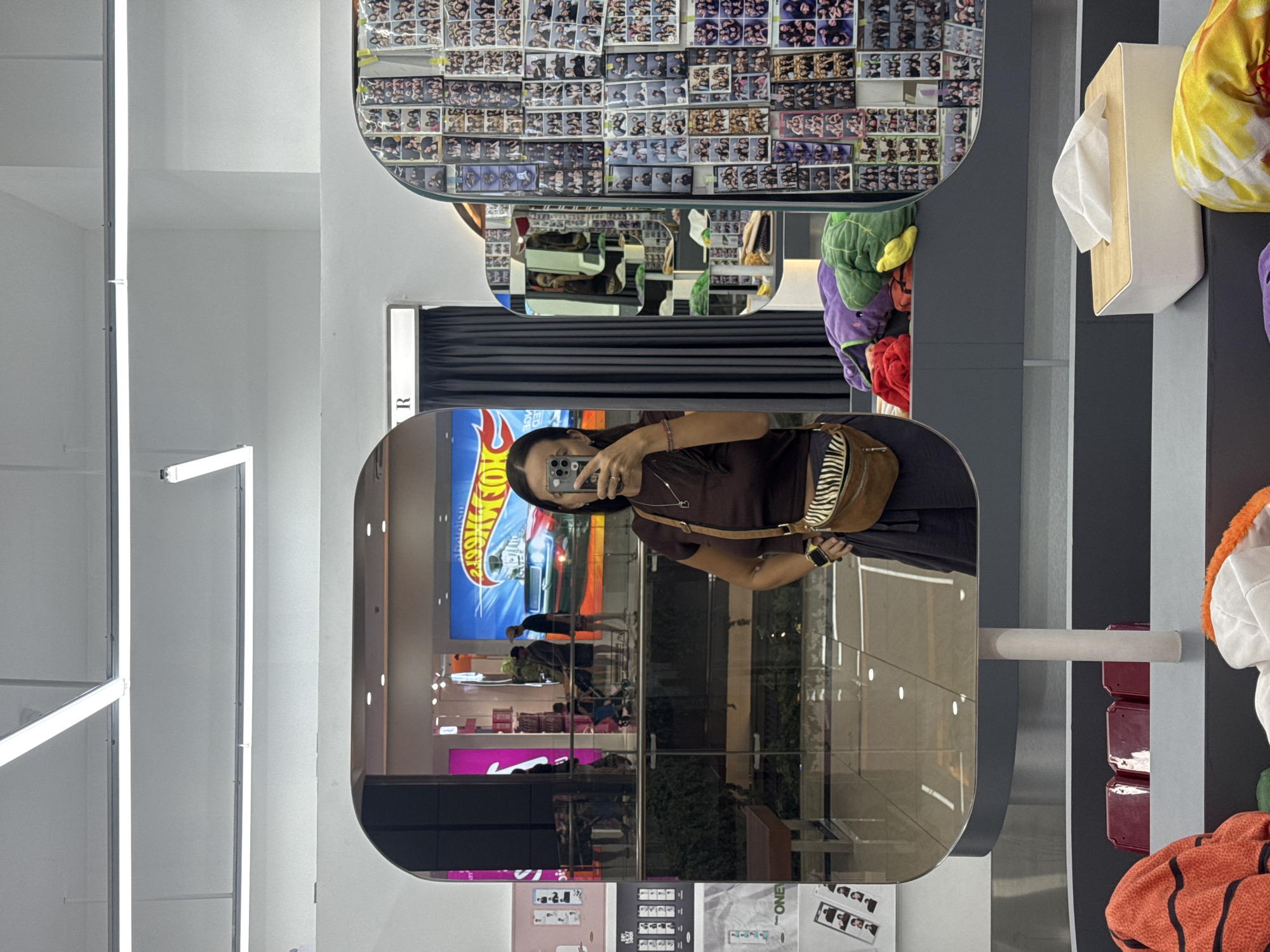A woman takes a mirror selfie in a well-lit retail store. She is wearing a dark brown t-shirt and a dark skirt, with a brown and white zebra-patterned crossbody bag. The store appears to be a clothing or novelty shop, with racks of clothing and displays of merchandise visible in the reflection. The background shows a large screen displaying a Hot Wheels advertisement, and several walls adorned with photo strips. In the foreground, there are folded blankets and a tissue box. The scene is clean and organized, with modern lighting fixtures and smooth, reflective surfaces. The store's location in Kuta, Indonesia is indicated by the signage visible in the reflection.