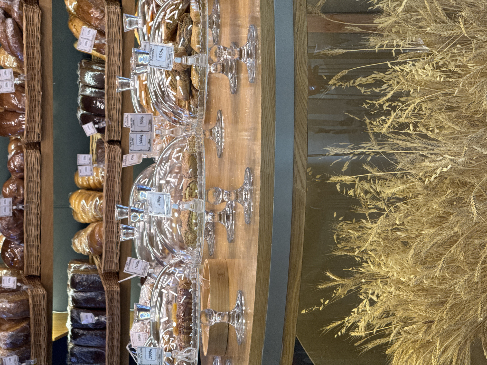 Here is a description of the image:
The image shows a display of baked goods, likely in a bakery or market. On the left, various loaves of bread are arranged in baskets on shelves. To the right, there are pastries or cookies displayed under glass domes on stands. Price tags are visible near the food items, with prices listed in numerals followed by what appears to be a Cyrillic abbreviation or symbol. Next to the bakery items, there are bushels of golden wheat, possibly used as a decorative element.