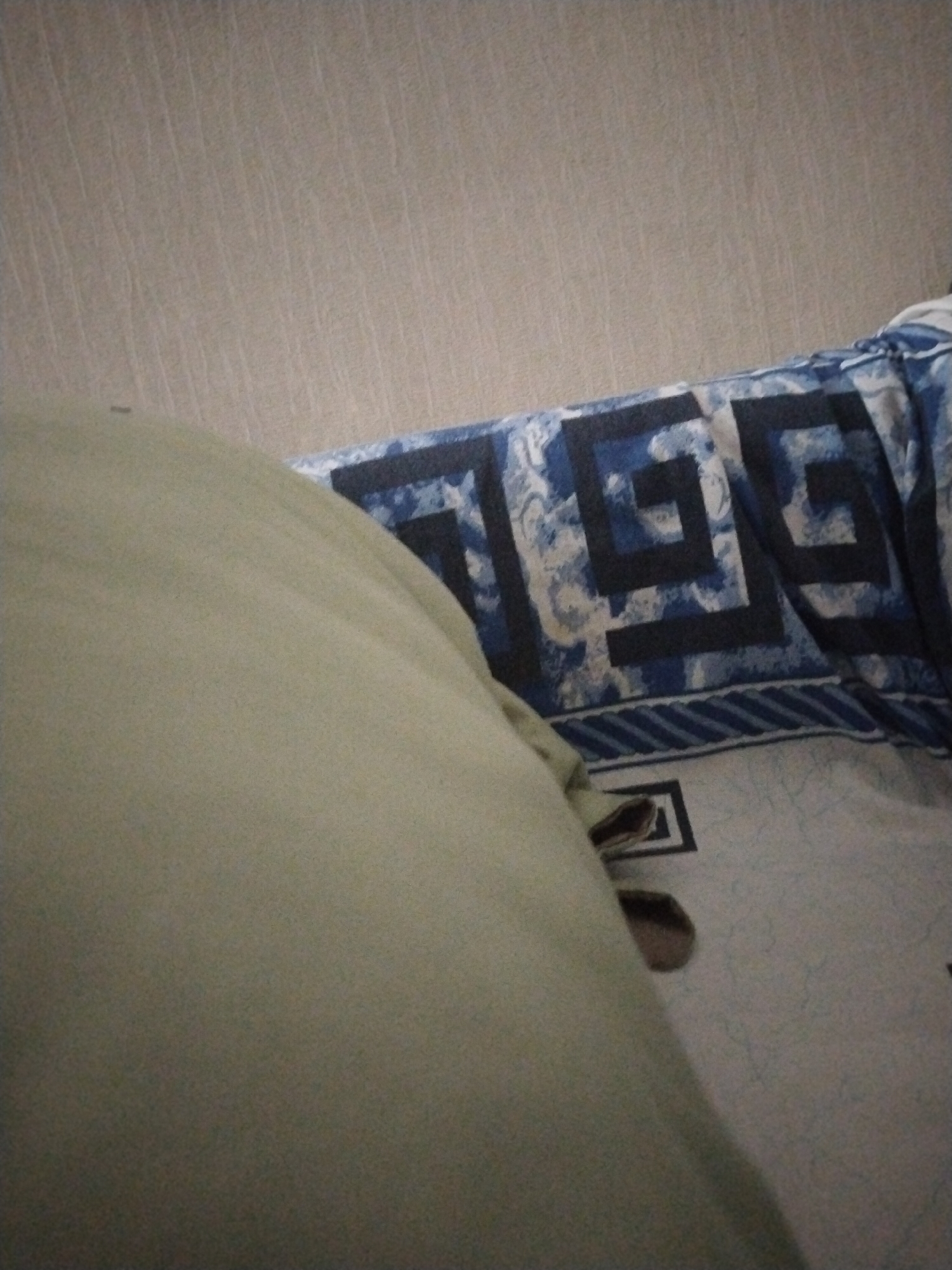 The image shows a close-up view of a bed with light-colored bedding in the foreground. A pillow with a light green or beige cover takes up the left side of the frame, obscuring much of the bed. In the background, a blanket or duvet features a geometric pattern in shades of blue and white, with repeating square-like designs. The pattern appears to be a maze or meander design. The setting seems to be indoors, likely a bedroom, given the presence of bedding. The lighting is dim, suggesting it might be evening or nighttime, or the room has low ambient light. There is no visible text or indication of any specific activity taking place, other than what appears to be the covered portion of a bed. The overall impression is a calm and subdued atmosphere. The location context of Ötegen batyr, Kazakhstan, does not provide any further visual clues within the image itself.