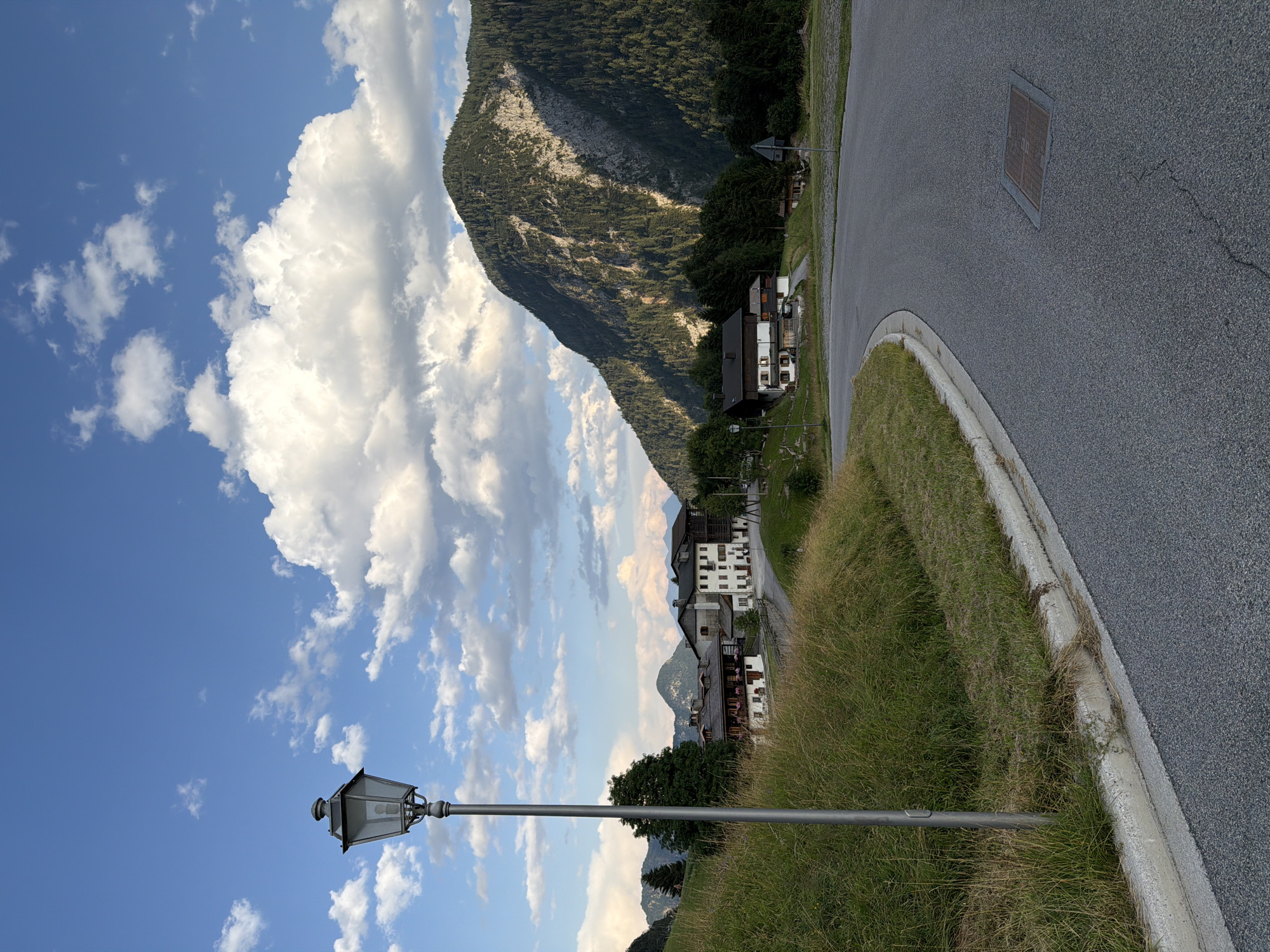 This is an outdoor, eye-level, full shot of a paved road curving through a mountainous landscape with scattered buildings and trees. The sky is blue with scattered white, fluffy clouds. In the foreground, a grey lamppost stands on the left side of the road, next to a grassy embankment. The road itself is dark grey asphalt. In the midground, several traditional alpine houses with wooden elements and balconies are visible, nestled among trees. The background is dominated by a large, steep mountain covered in dense green pine trees, with patches of rocky grey exposed on its upper slope. The lighting suggests it is either late afternoon or early morning, with shadows cast by the mountain onto its slopes and the surrounding trees. The location is the municipality of Sauris / Zahre, Italy, known for its alpine setting and distinct culture. There are no people visible in the image.