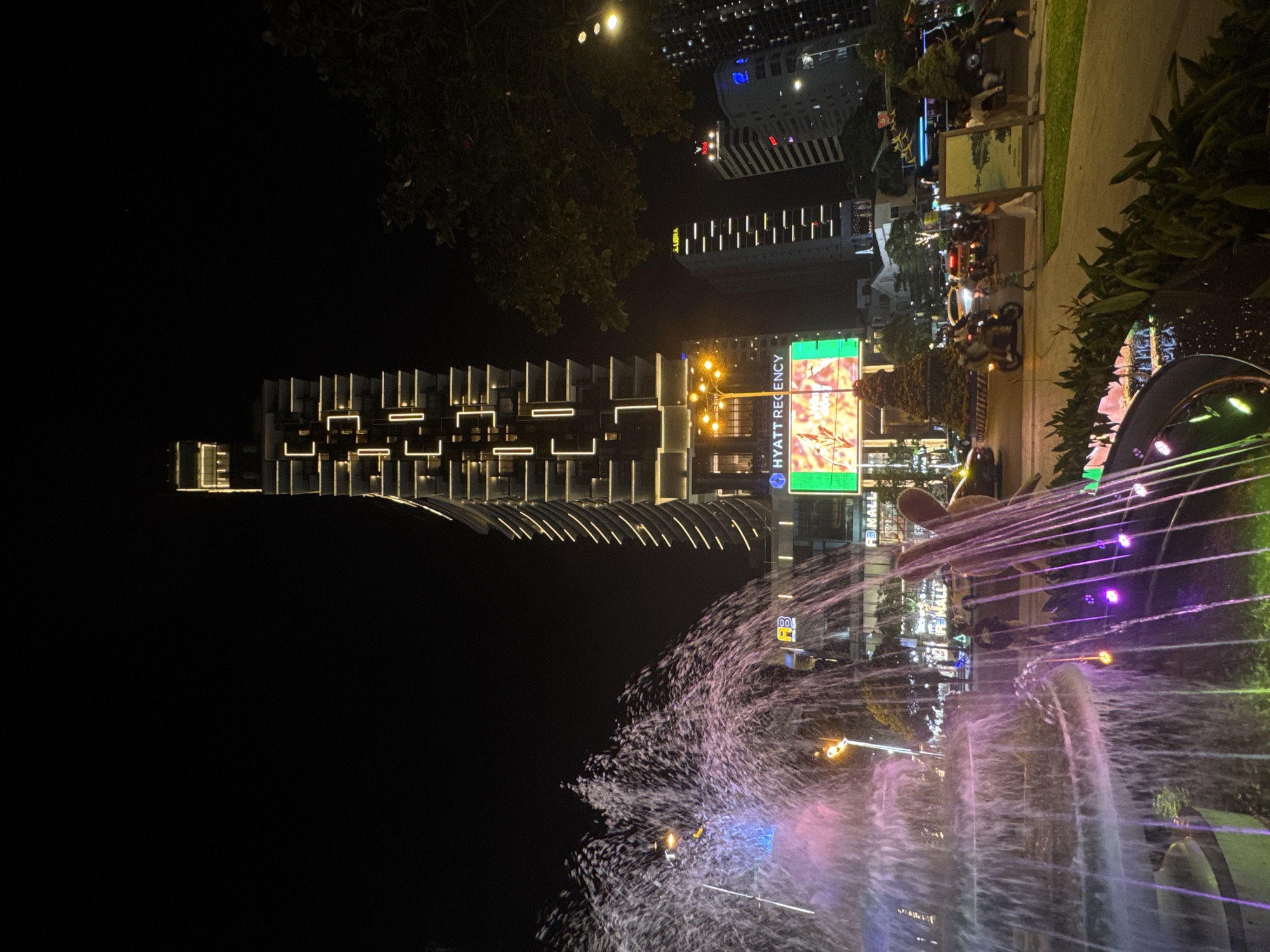 At night, a fountain with pink and purple lighting sprays water upwards in the foreground. Behind the fountain, streetlights illuminate a sidewalk with people, motorbikes, and artwork. The Hyatt Regency building is visible further back, along with other tall buildings with bright lights. The image suggests an urban environment, specifically near the Hyatt Regency in an unidentified city, possibly in Vietnam, due to the presence of motorbikes and street signs.