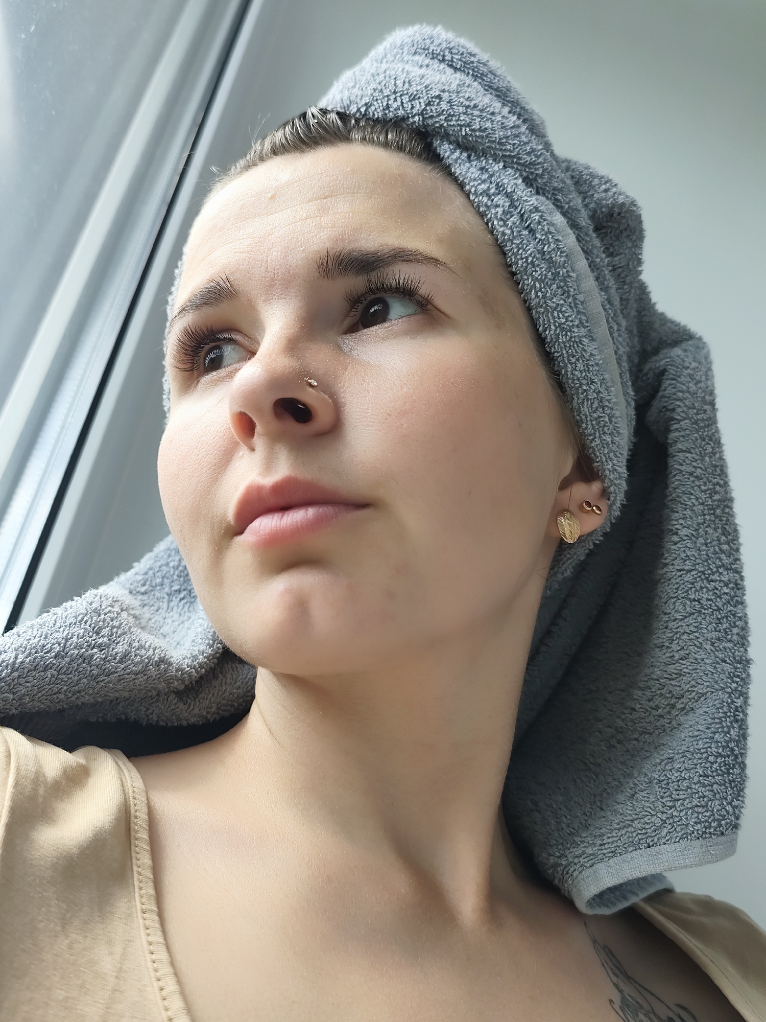 A close-up shot captures a fair-skinned person with wet hair wrapped in a gray towel. The person has a nose piercing and gold-colored earrings. They gaze towards a window. Their attire consists of a light tan or beige top. A partial view reveals a tattoo on their upper chest. The location is indicated as Фоки, Russia, though specific details tying the image to a particular area within the city are absent.